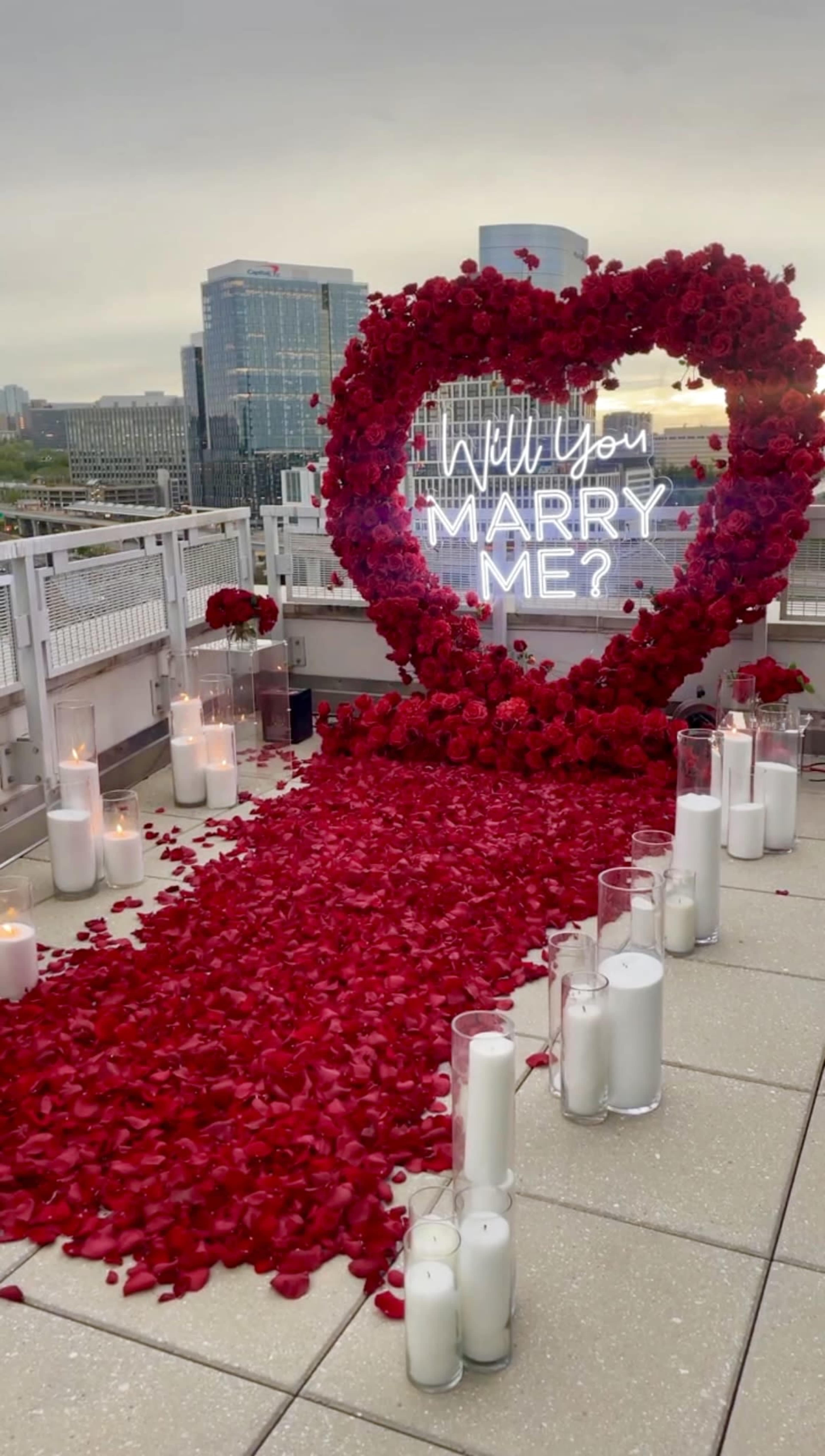 A heart-shaped floral arrangement surrounded by scattered rose petals and illuminated candles decorates a rooftop space for a marriage proposal.