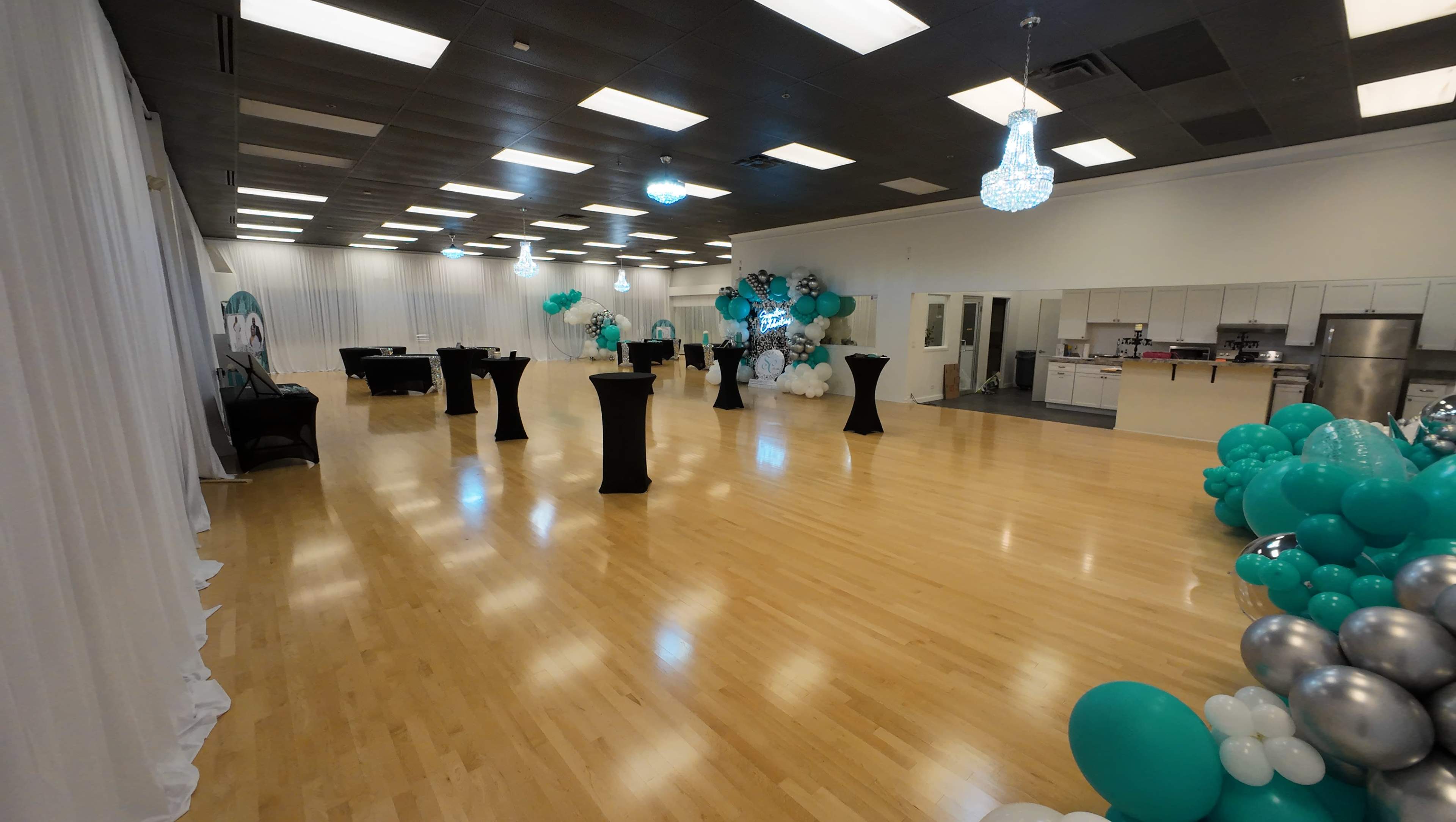 An empty event space featuring a wooden floor, decorated with black tables, balloon arrangements, and a large white curtain backdrop.