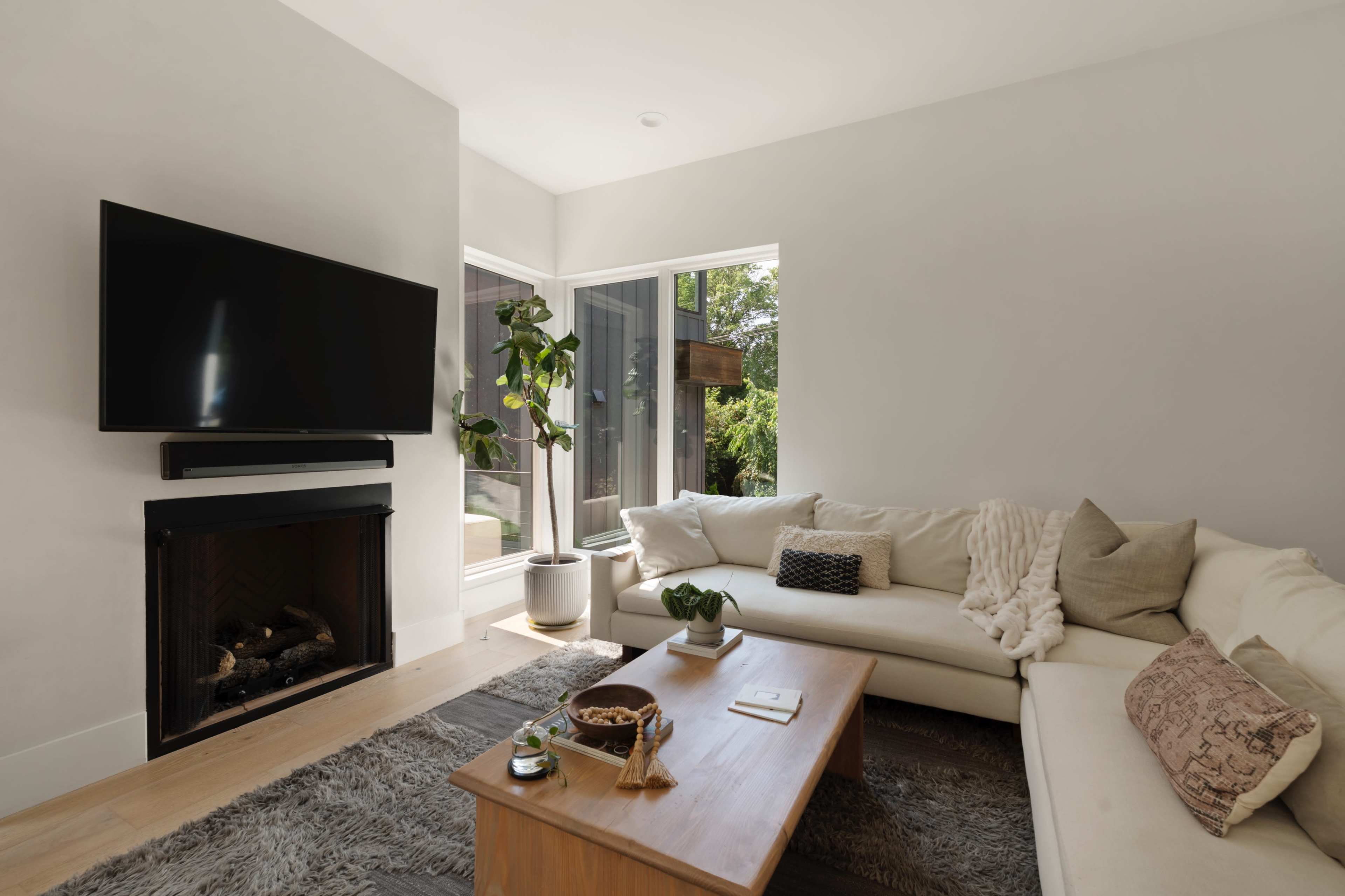 A modern living room features a large sofa, a wooden coffee table, a television mounted on the wall, and large windows overlooking a garden.