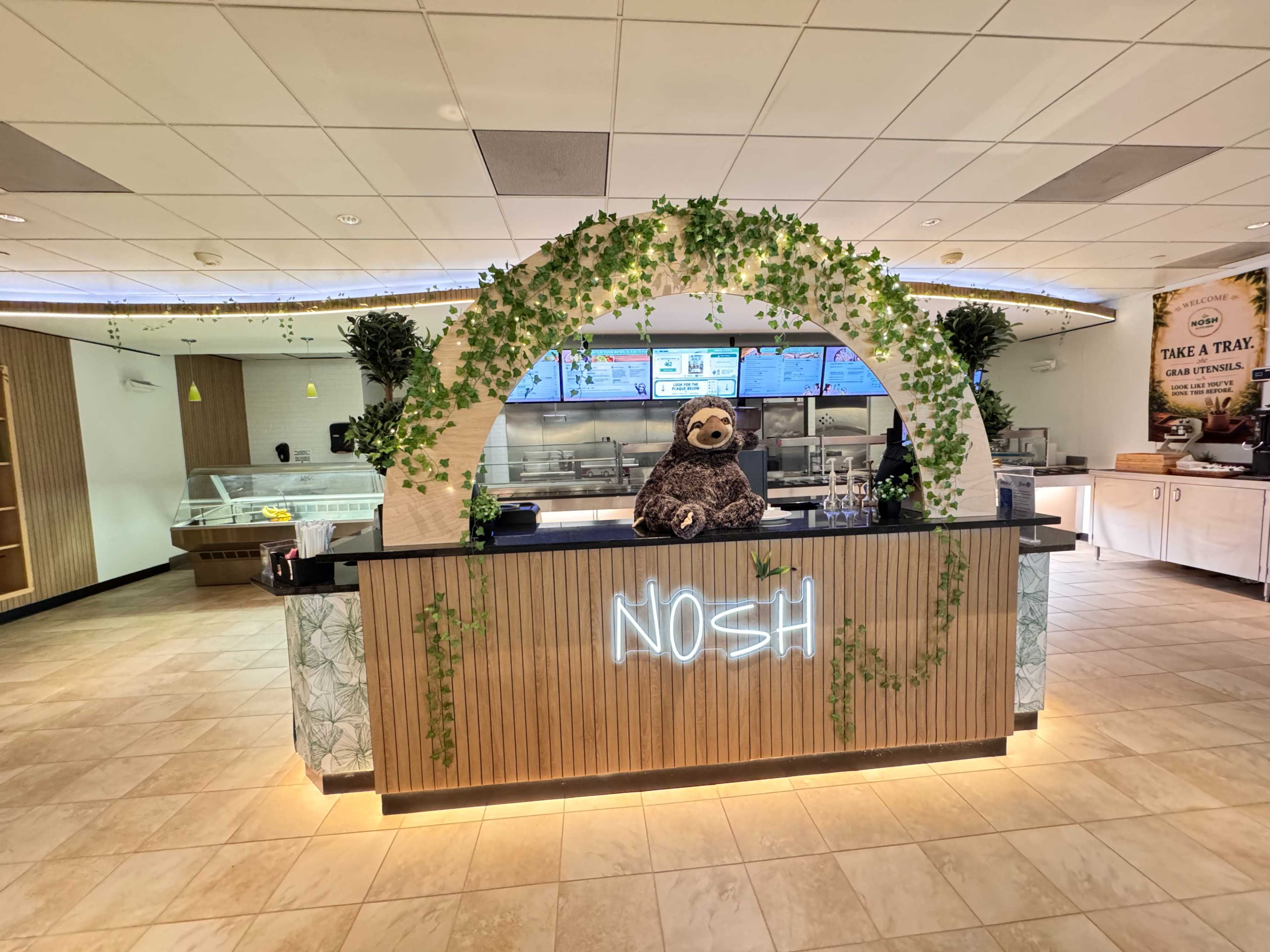 The image shows a café counter with a neon sign reading "NOSH," adorned with greenery and featuring a stuffed sloth.