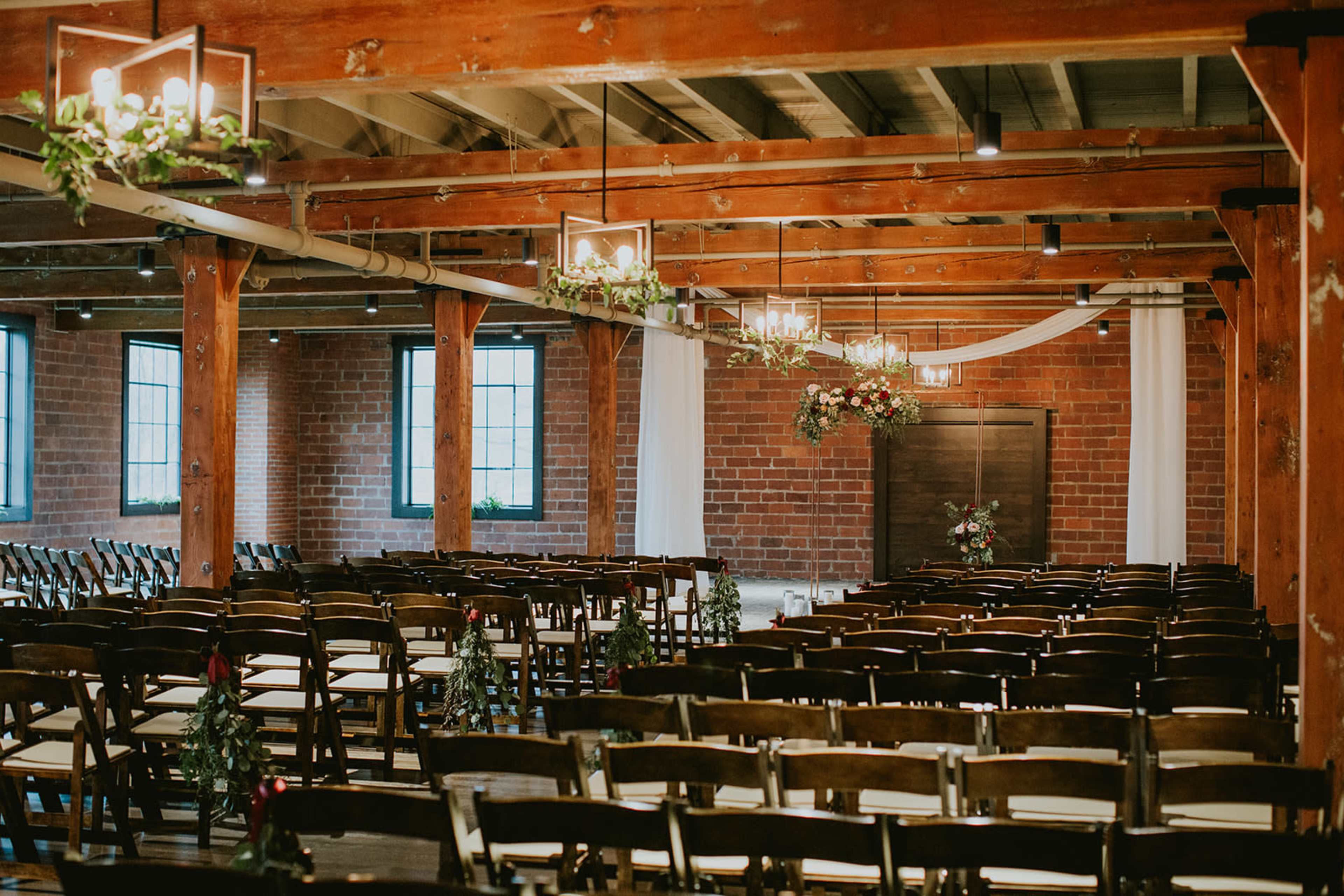 A wedding venue with rows of wooden chairs arranged facing a decorative backdrop and exposed brick walls, featuring hanging lights and greenery.