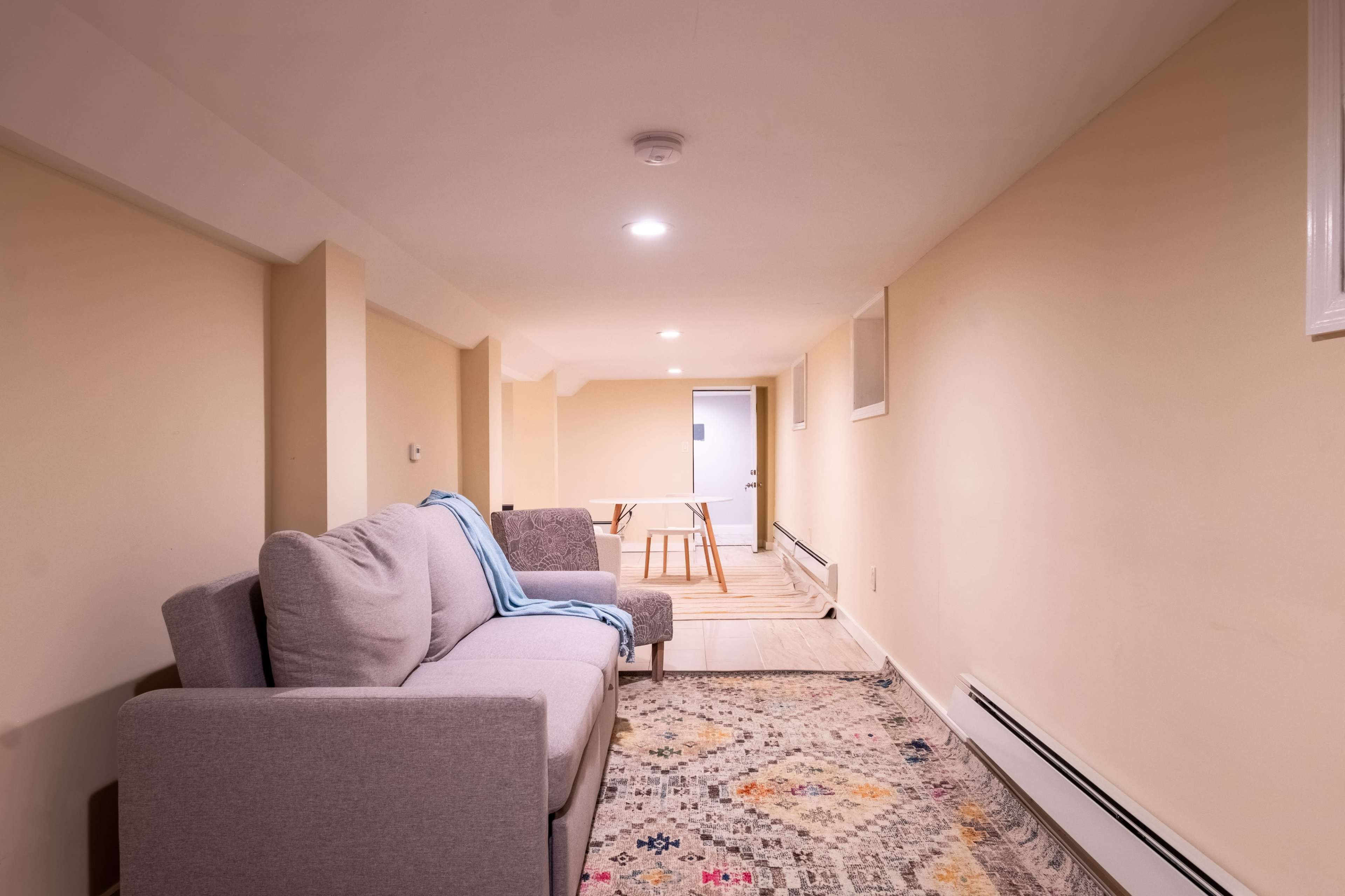 A narrow, brightly lit basement features a gray sofa with a blanket, a small table, and a patterned rug along the floor.