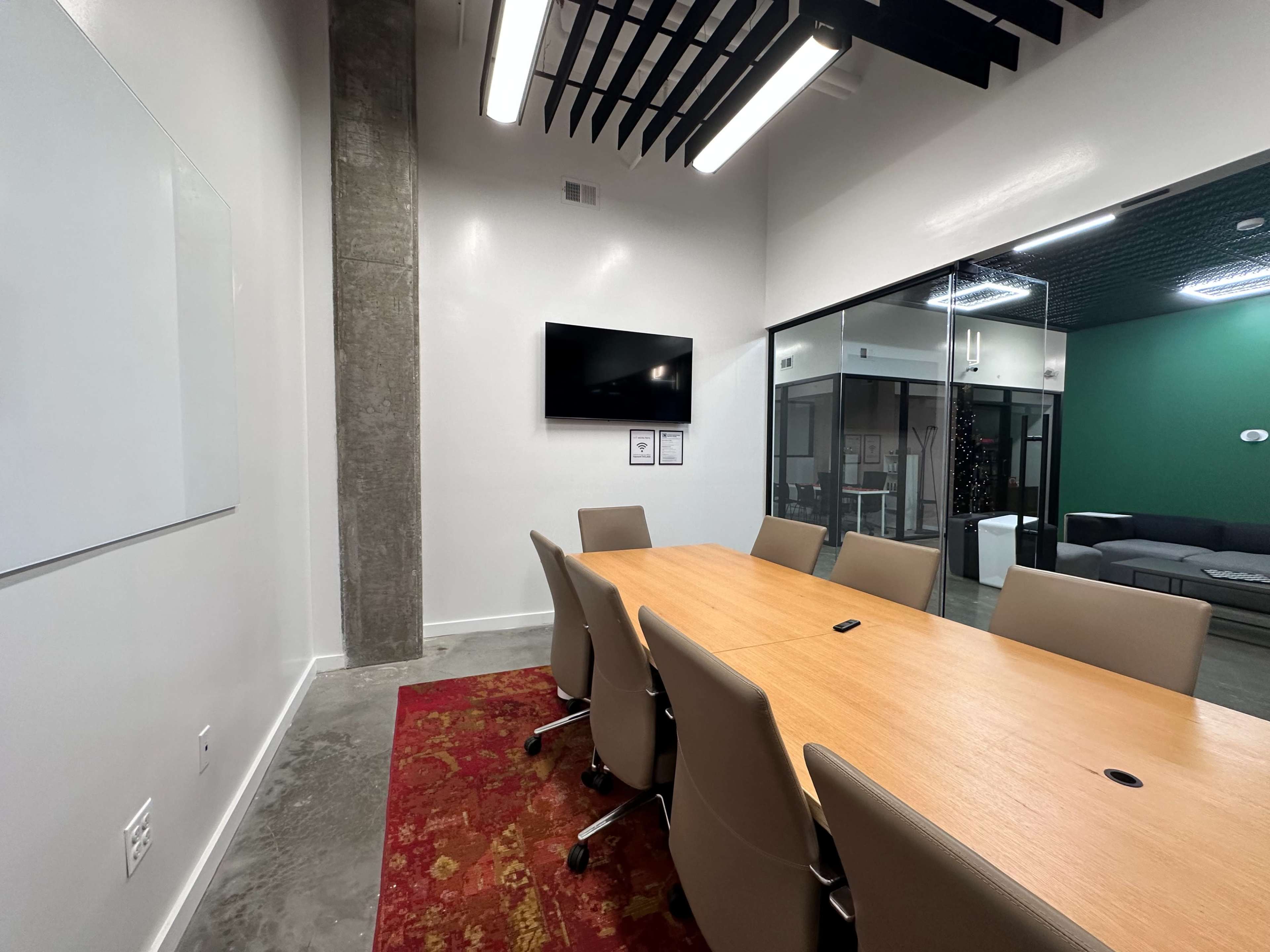 The image shows a modern conference room with a long wooden table, ergonomic chairs, a wall-mounted television, and glass walls overlooking a lounge area.
