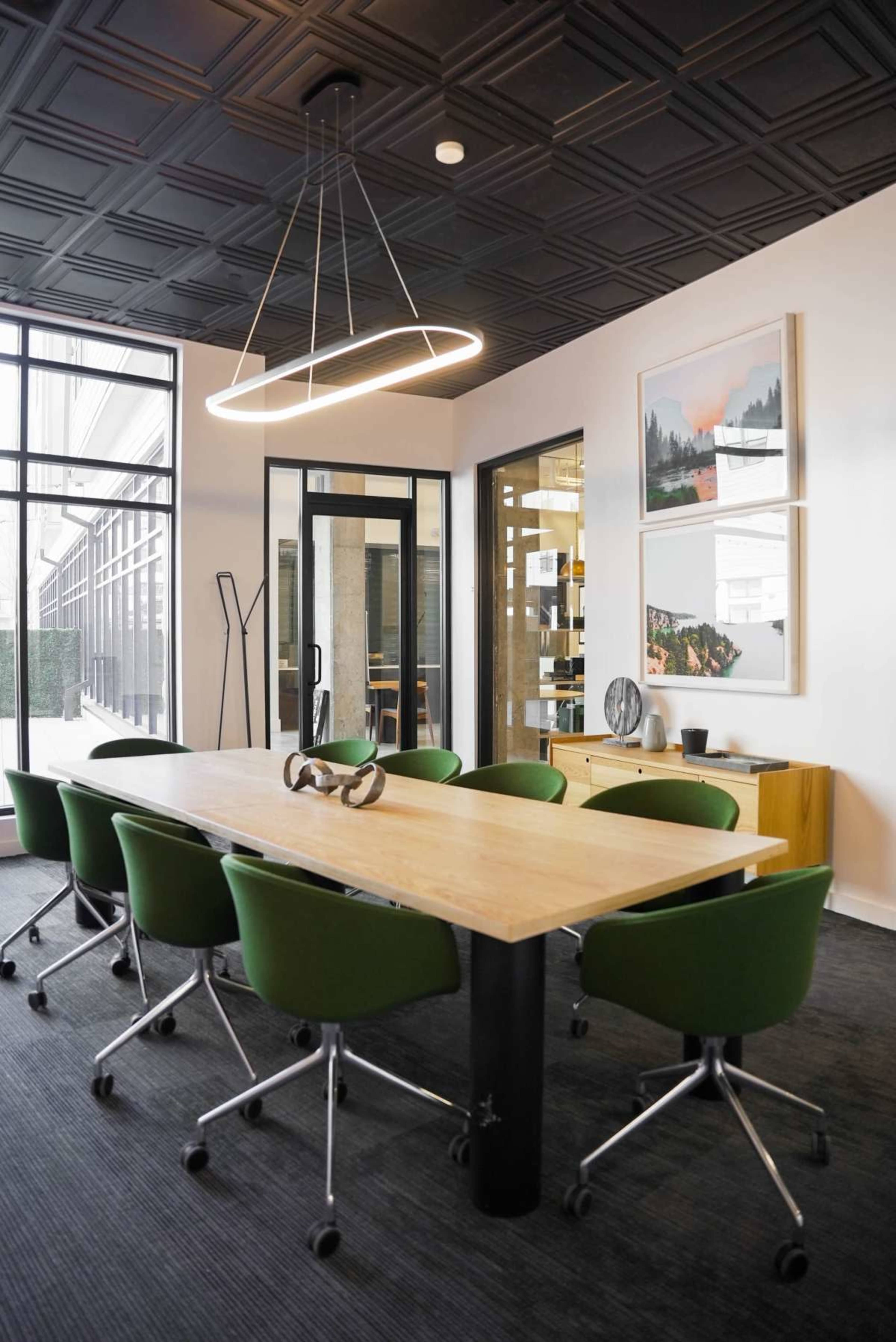 A modern conference room features a long wooden table surrounded by green rolling chairs, with a contemporary light fixture overhead and artwork on the walls.