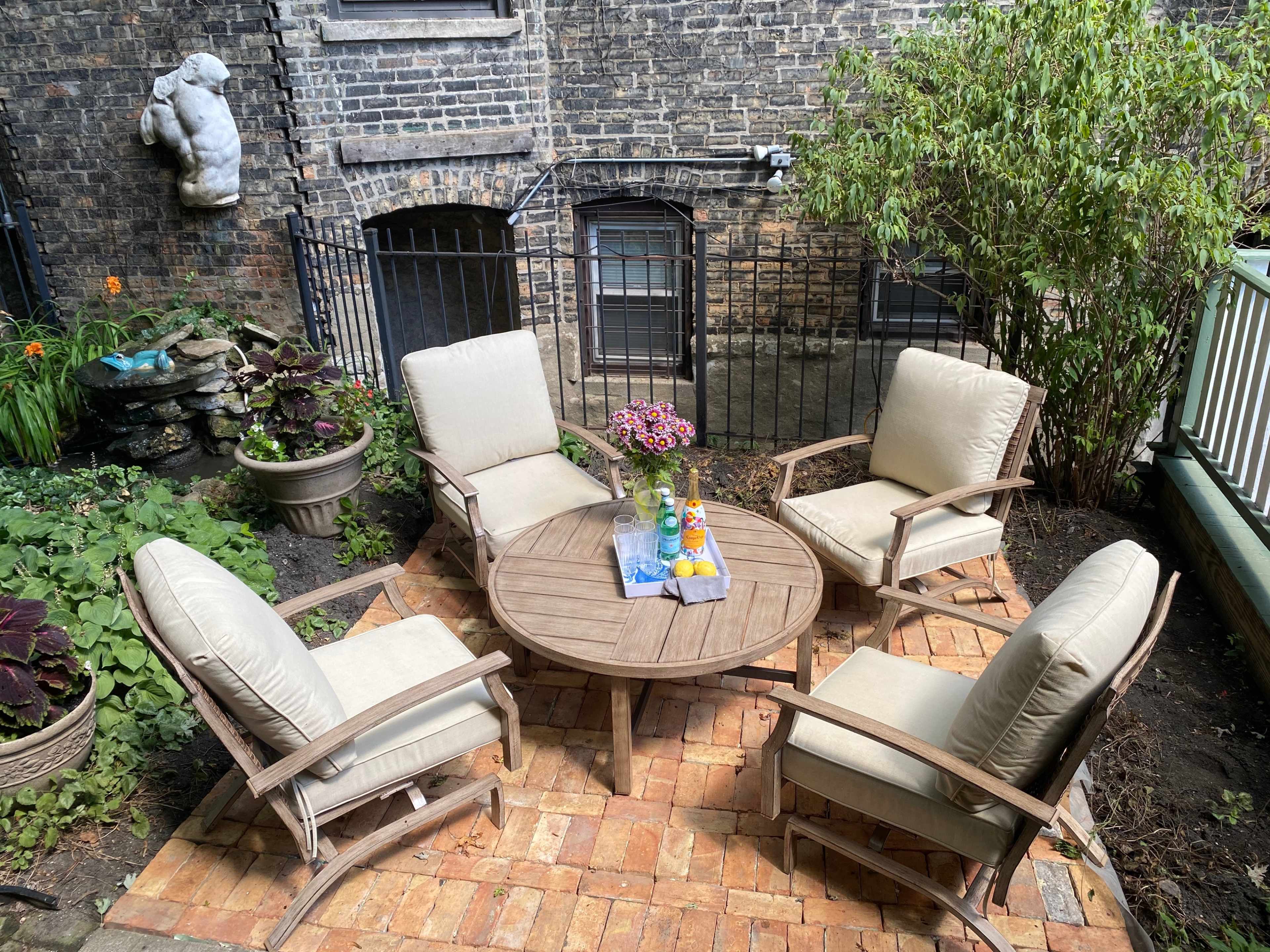 A circular wooden table surrounded by four cushioned chairs sits in a brick-paved outdoor space, with plants and a stone feature in the background.