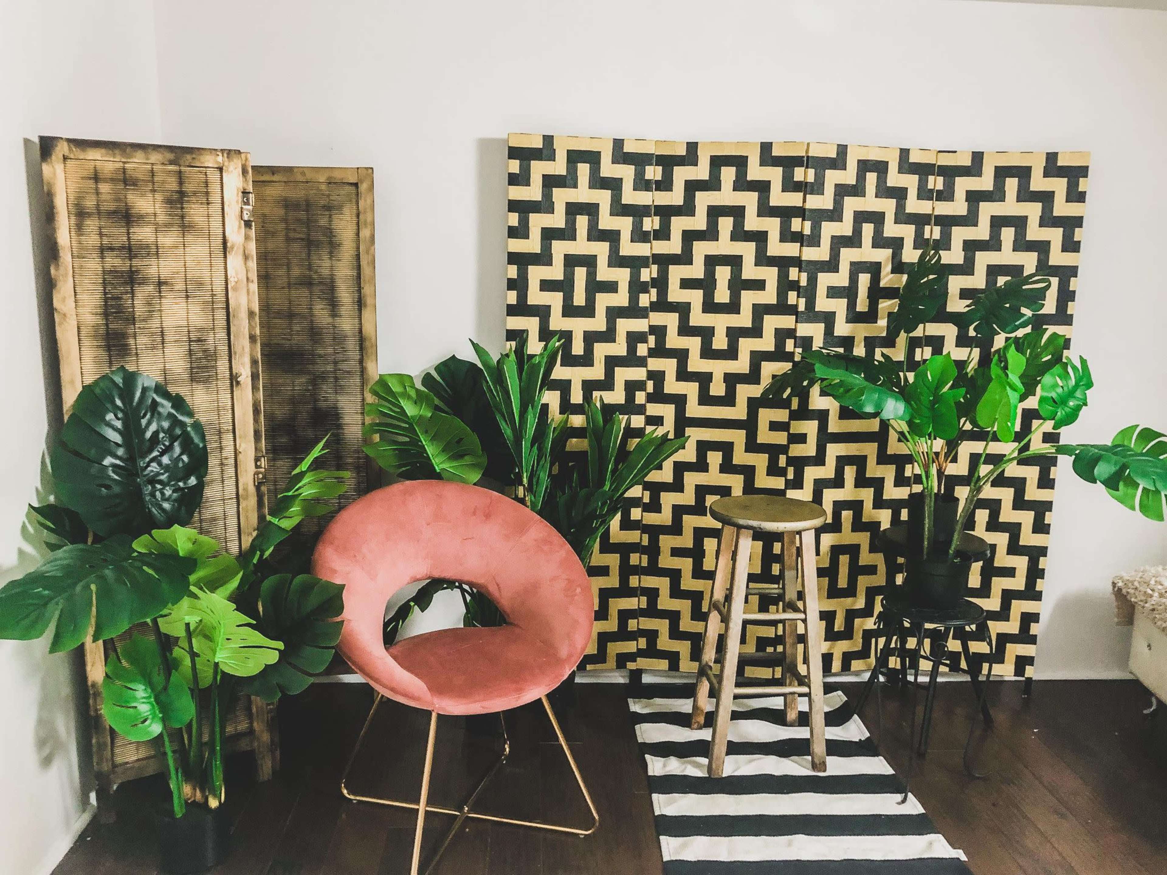 A modern room featuring a pink chair, a wooden stool, and various green plants against a patterned backdrop.