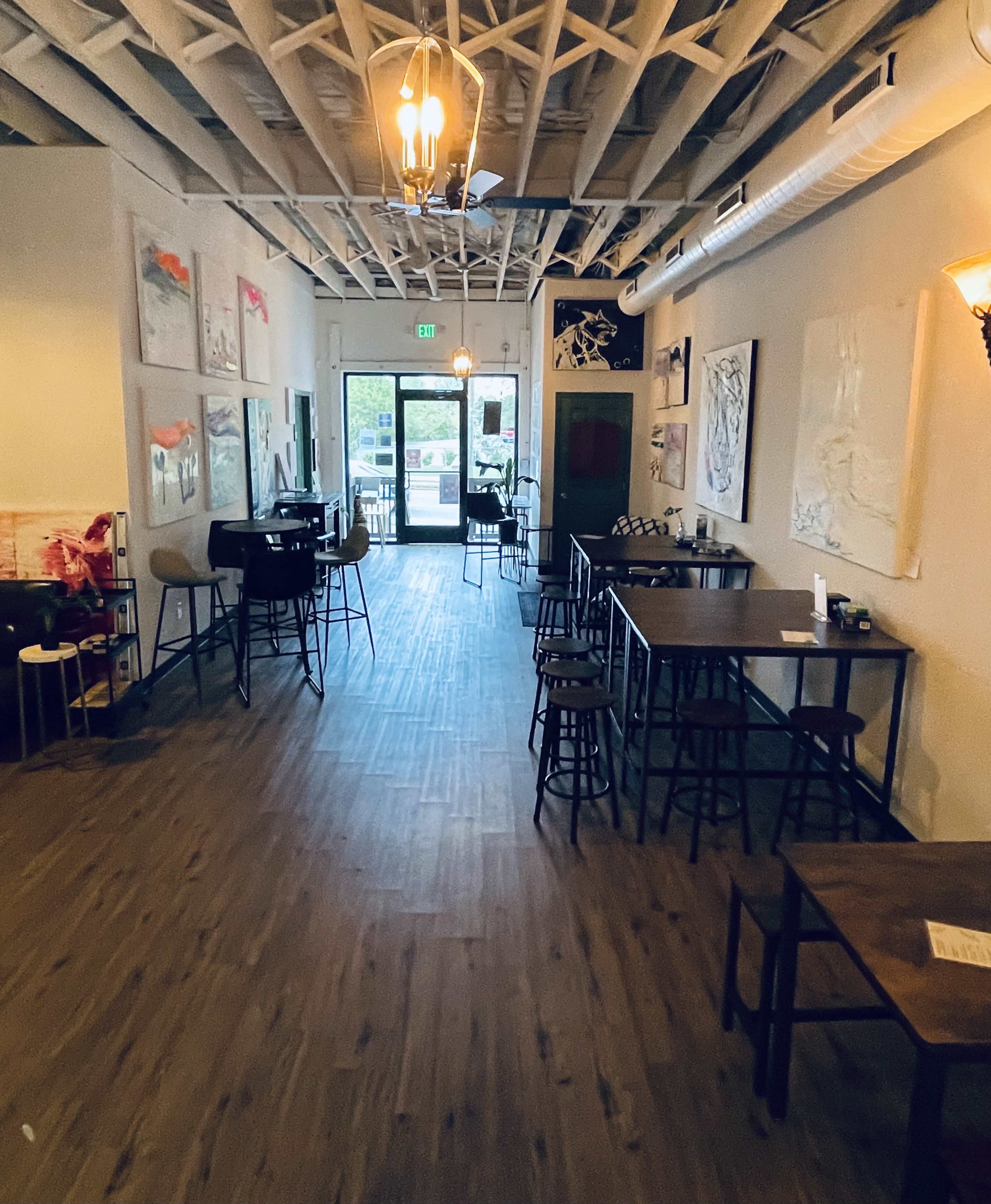 The image shows a spacious interior of a restaurant or café with wooden flooring, tables with chairs, and artwork displayed on the walls.