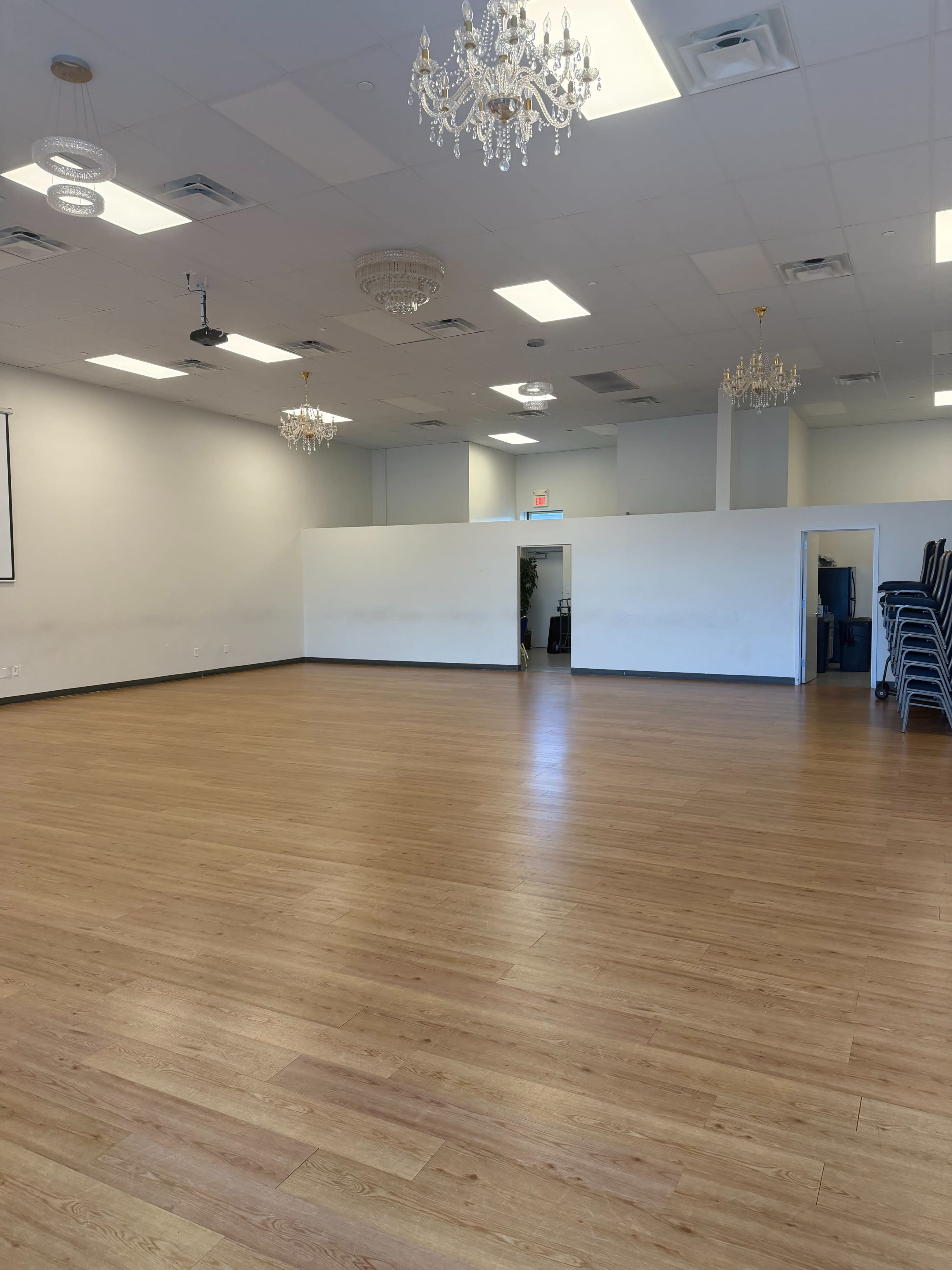 The image shows a spacious, empty room with wooden flooring, white walls, and several chandeliers hanging from the ceiling.