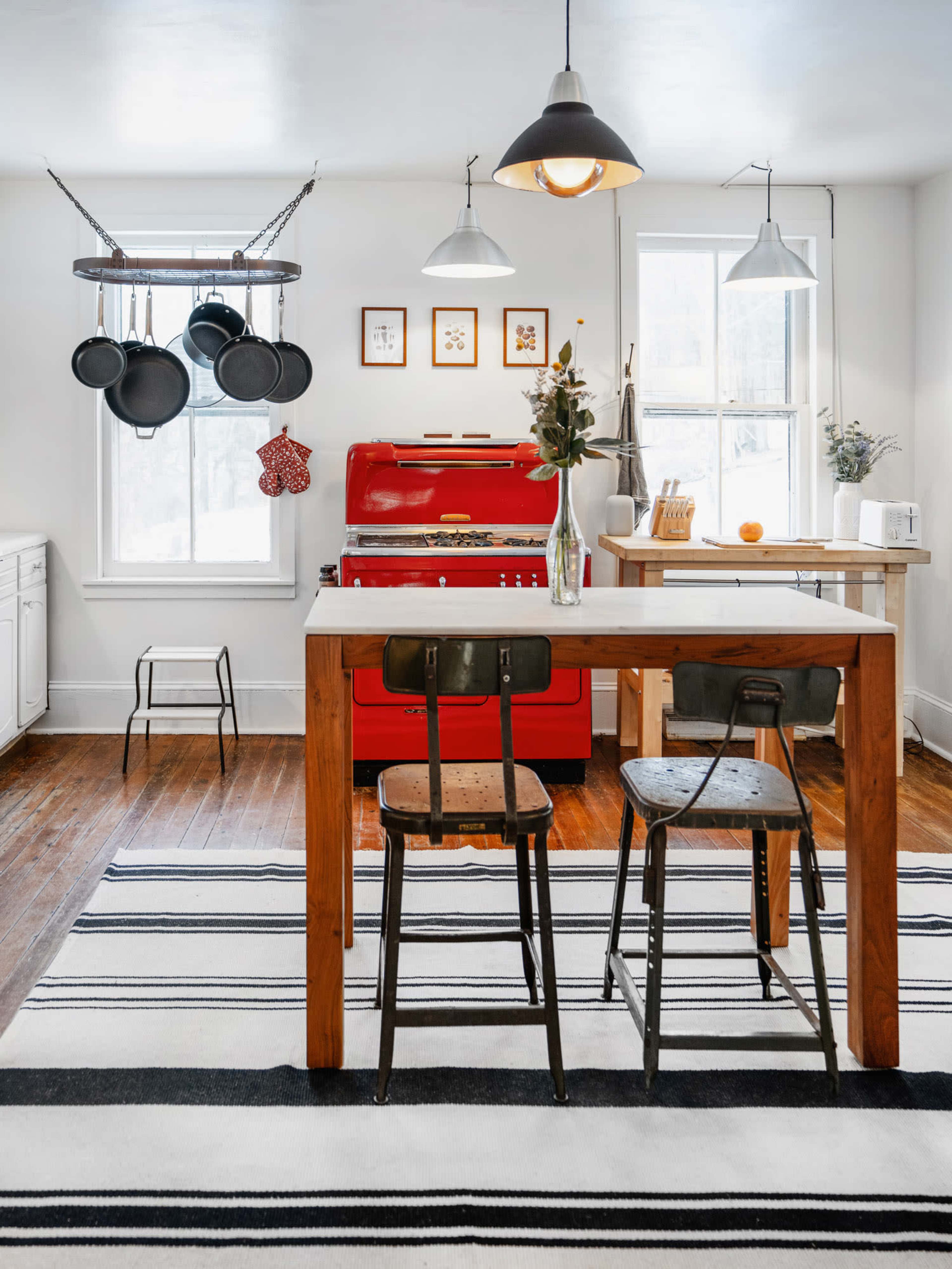A modern kitchen with a red stove, wooden island, two metal stools, and hanging pots, all set against a striped black and white rug.