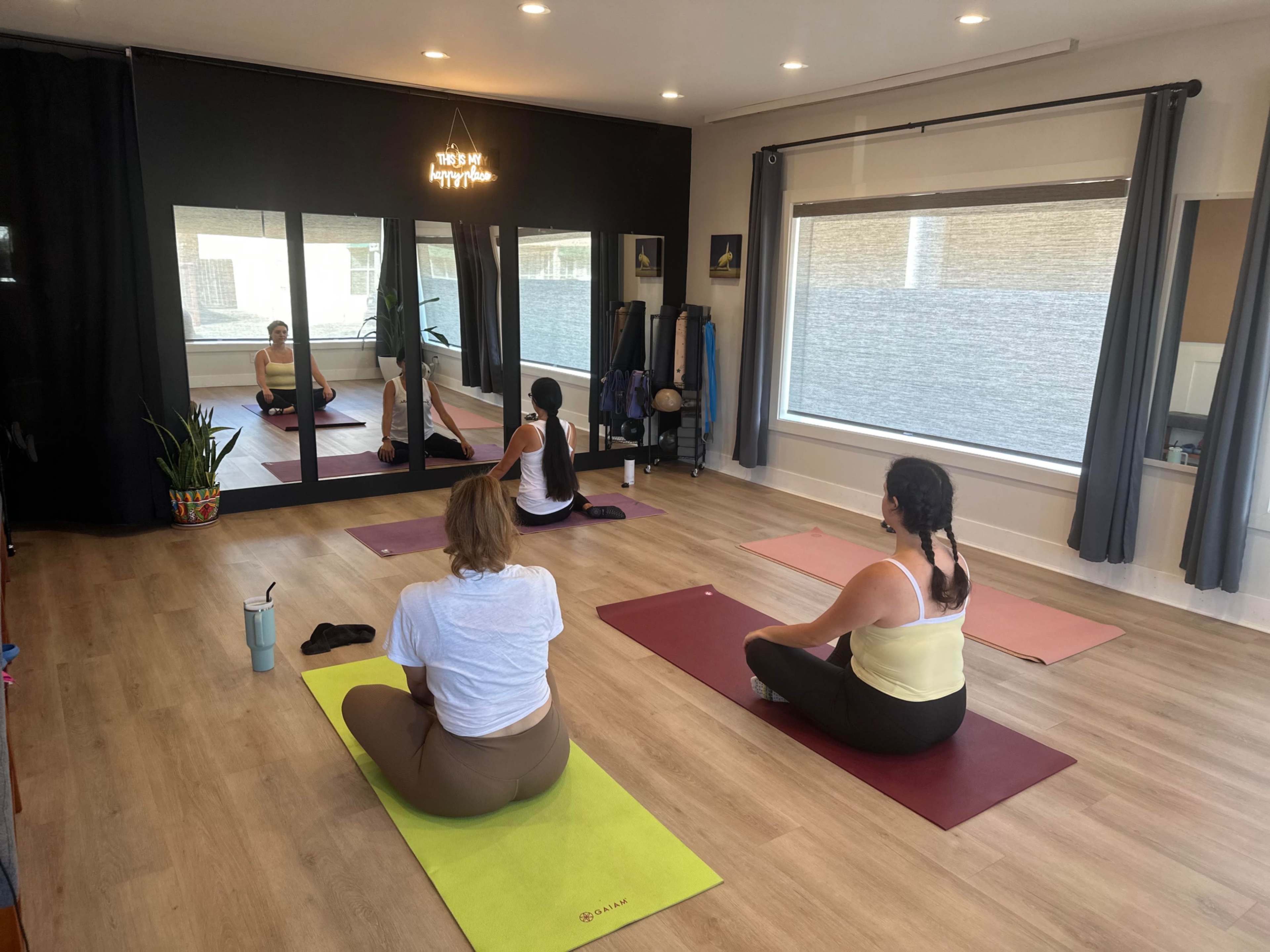 Three people are participating in a yoga session on mats in a brightly lit studio with mirrors and plants.