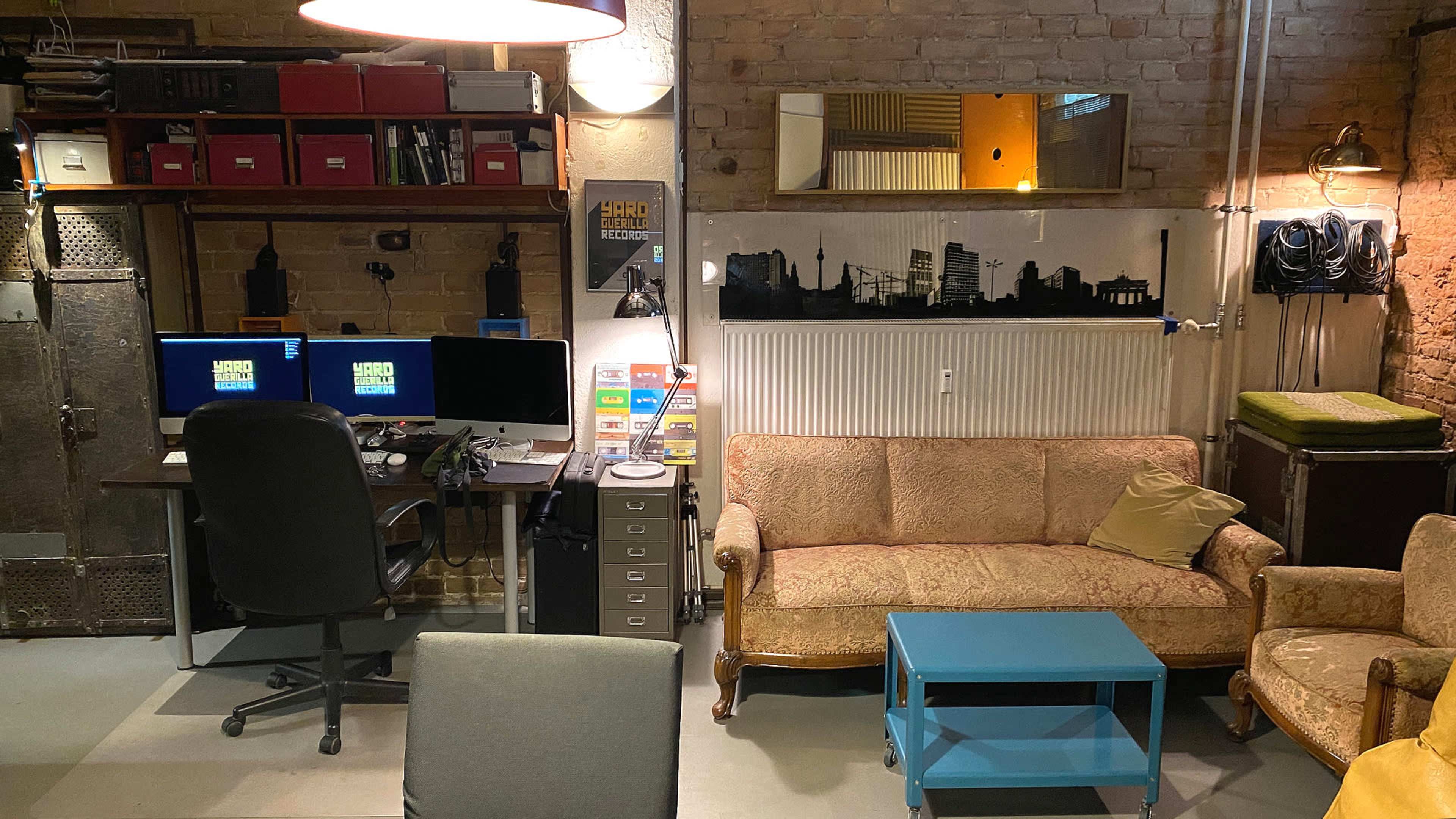 The image shows a room with a desk setup featuring dual monitors, a vintage sofa, and a blue coffee table, alongside exposed brick walls and various storage options.