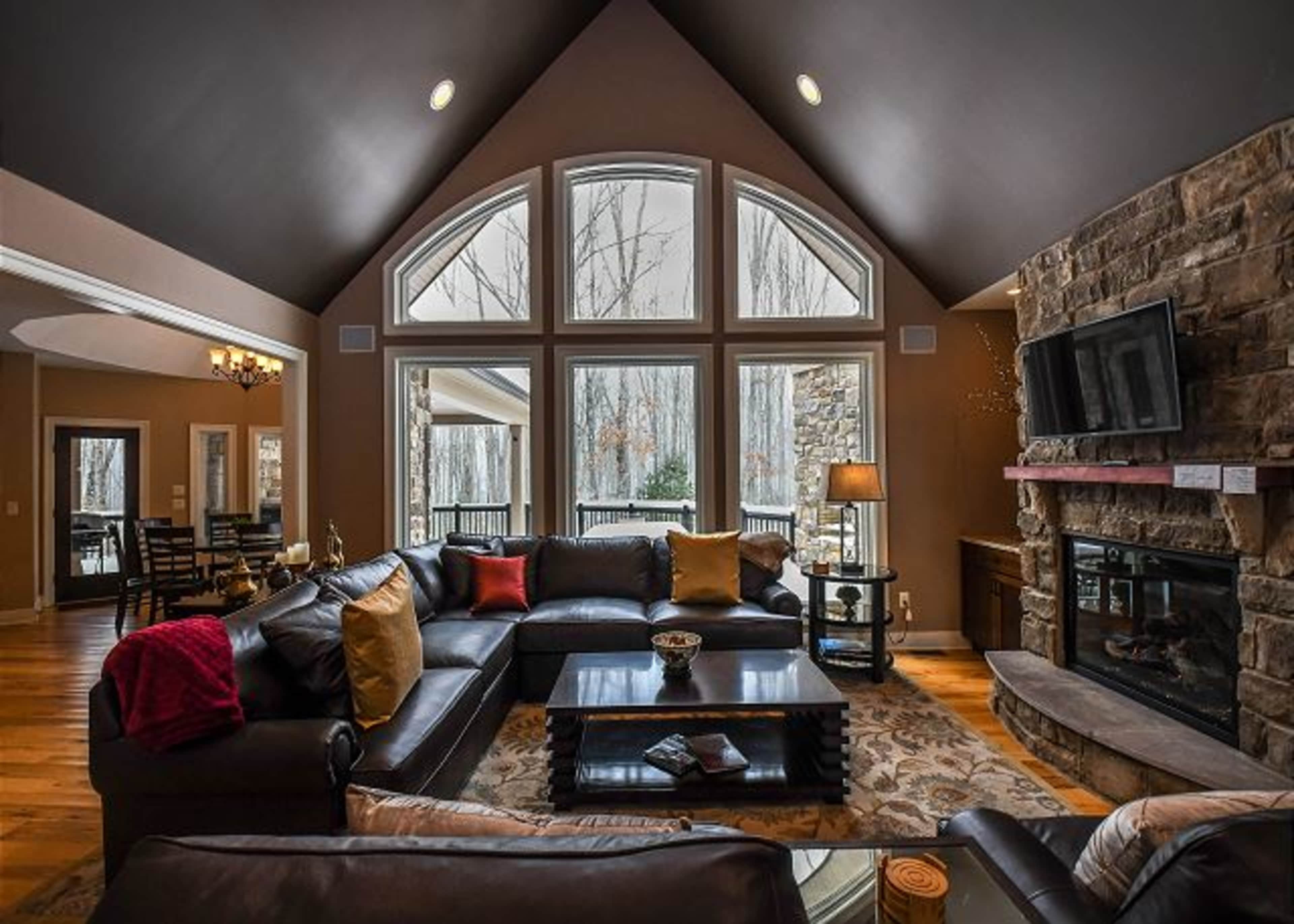 A modern living room features large arched windows, a stone fireplace, and a dark leather seating arrangement with a coffee table.