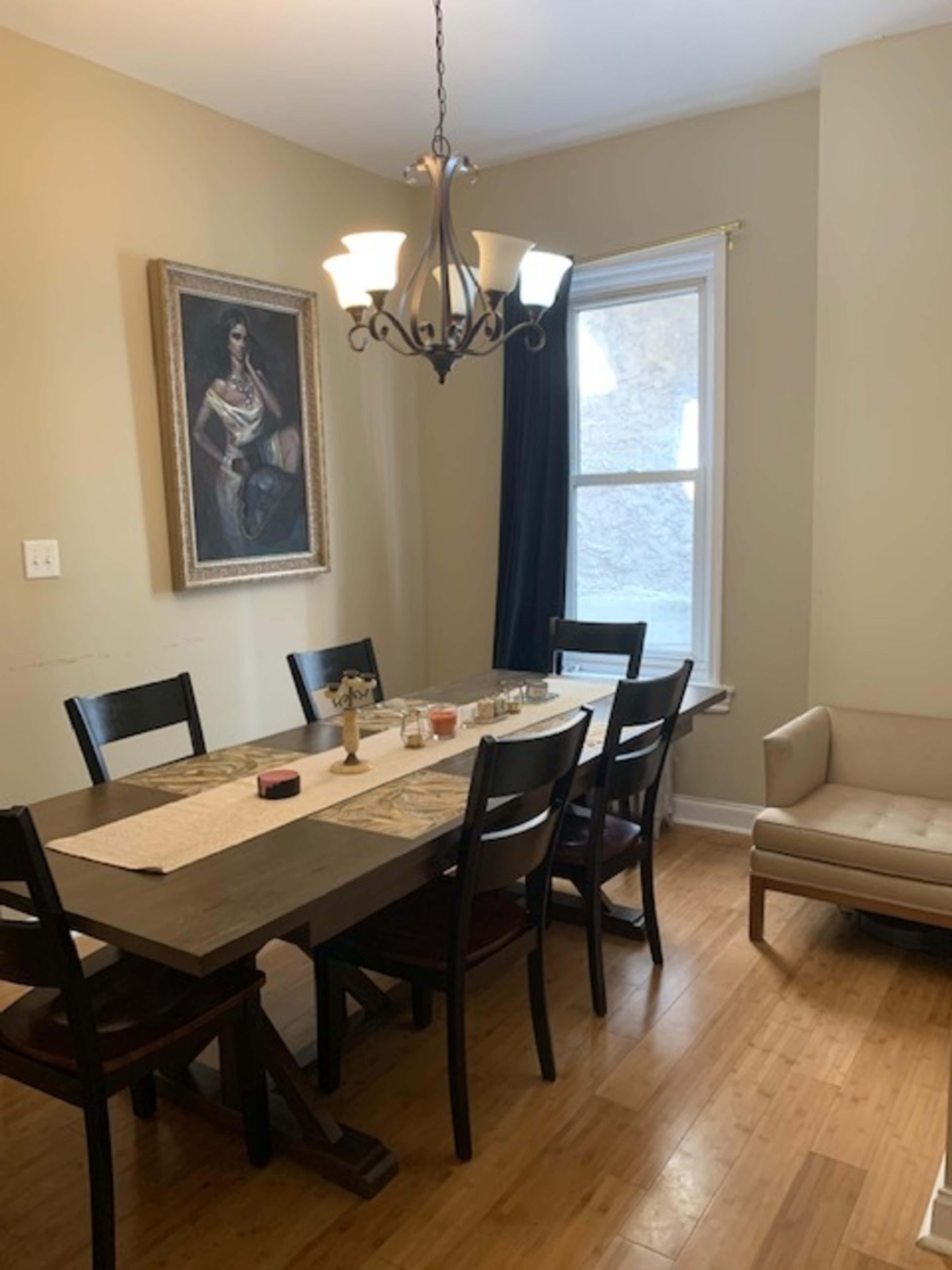 A dining room features a wooden table surrounded by eight chairs, a chandelier above, and a large portrait on the wall.
