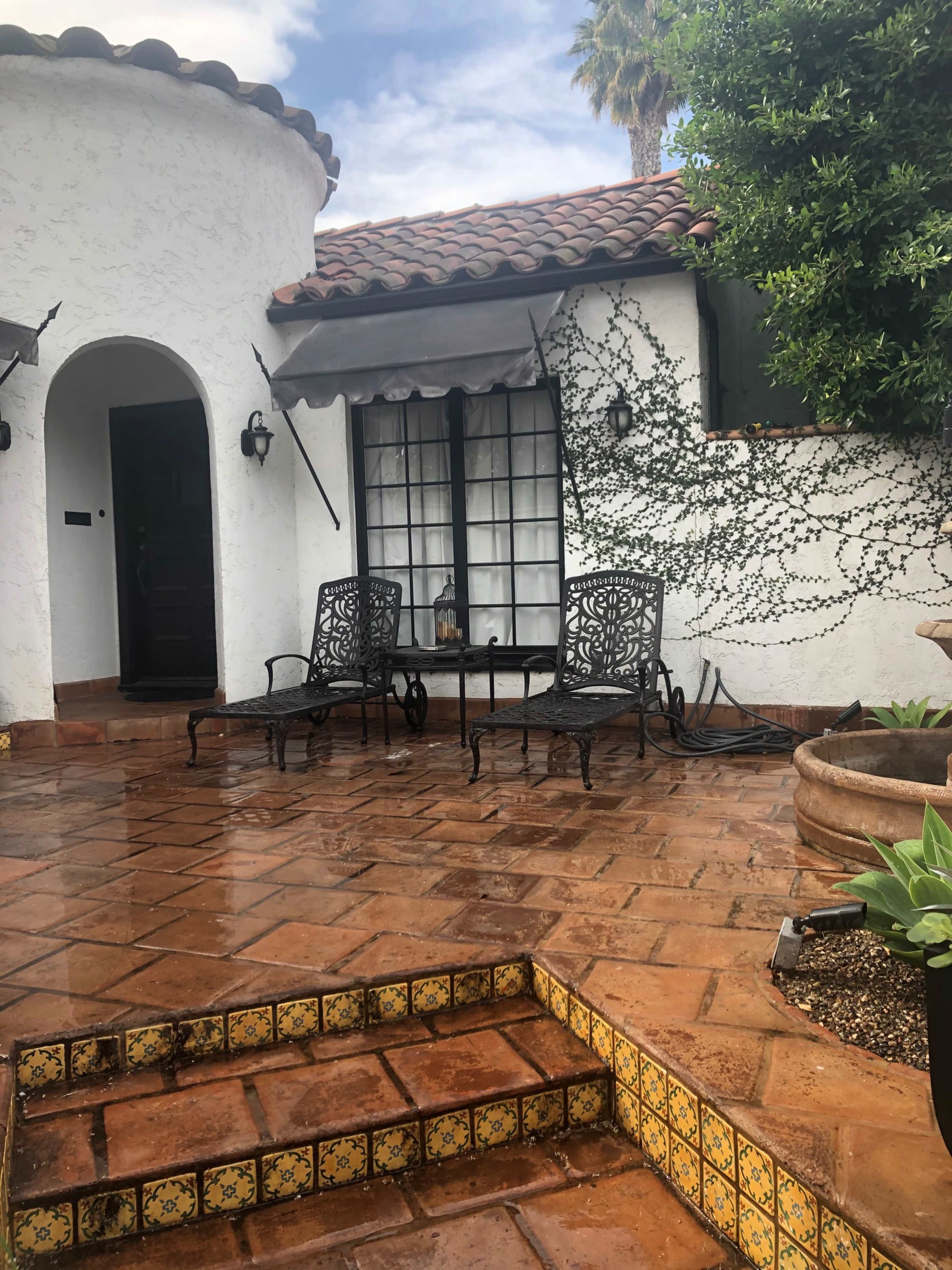 A patio area features two metal chairs beside a table, with decorative tile steps leading to a doorway and greenery climbing the wall.