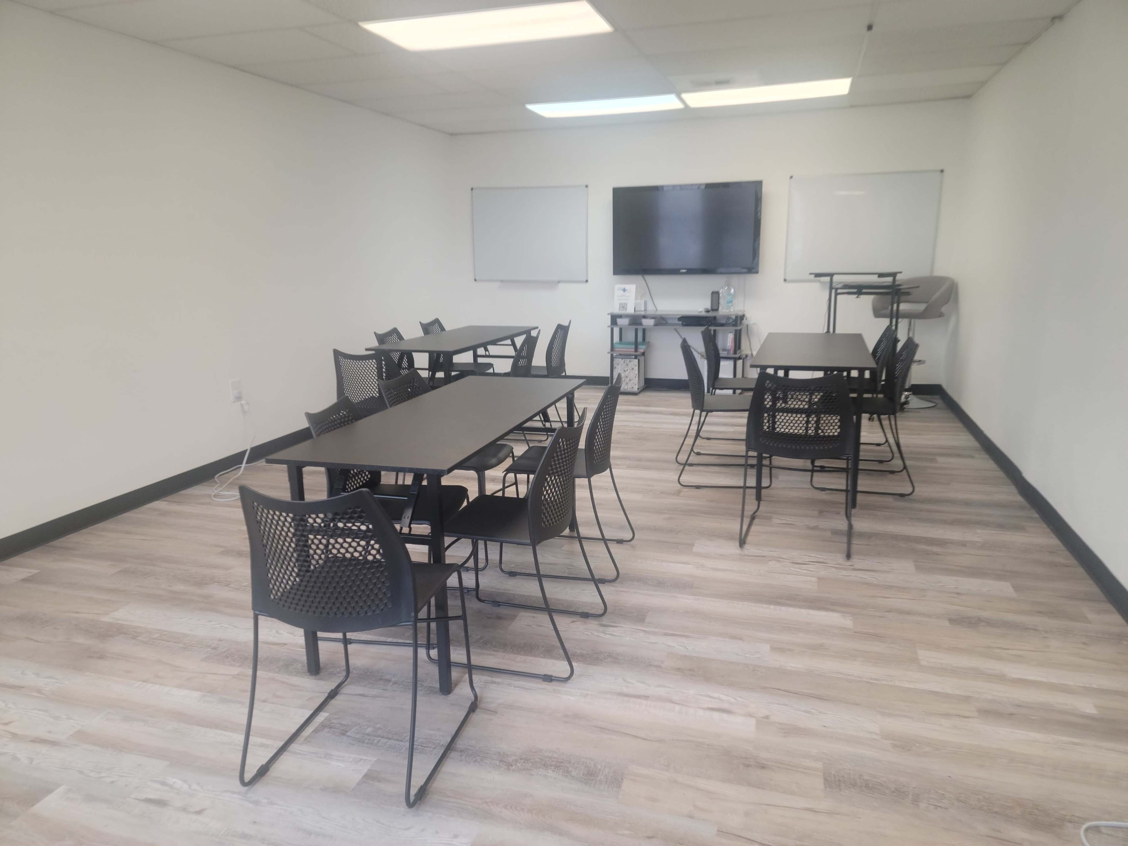 A classroom features several rectangular tables surrounded by black mesh chairs, with a television and whiteboards mounted on the walls.
