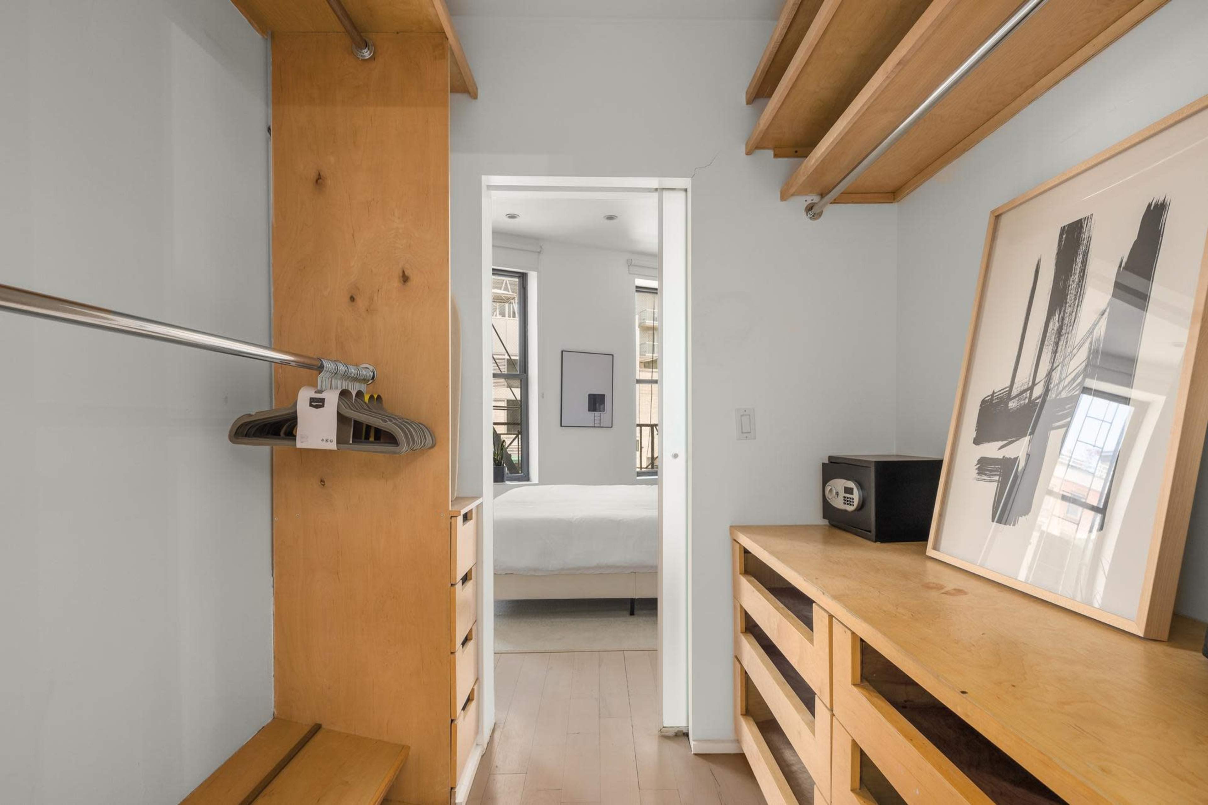 A small room with wooden shelves and a clothing rack, leading to a bedroom visible through an open door.