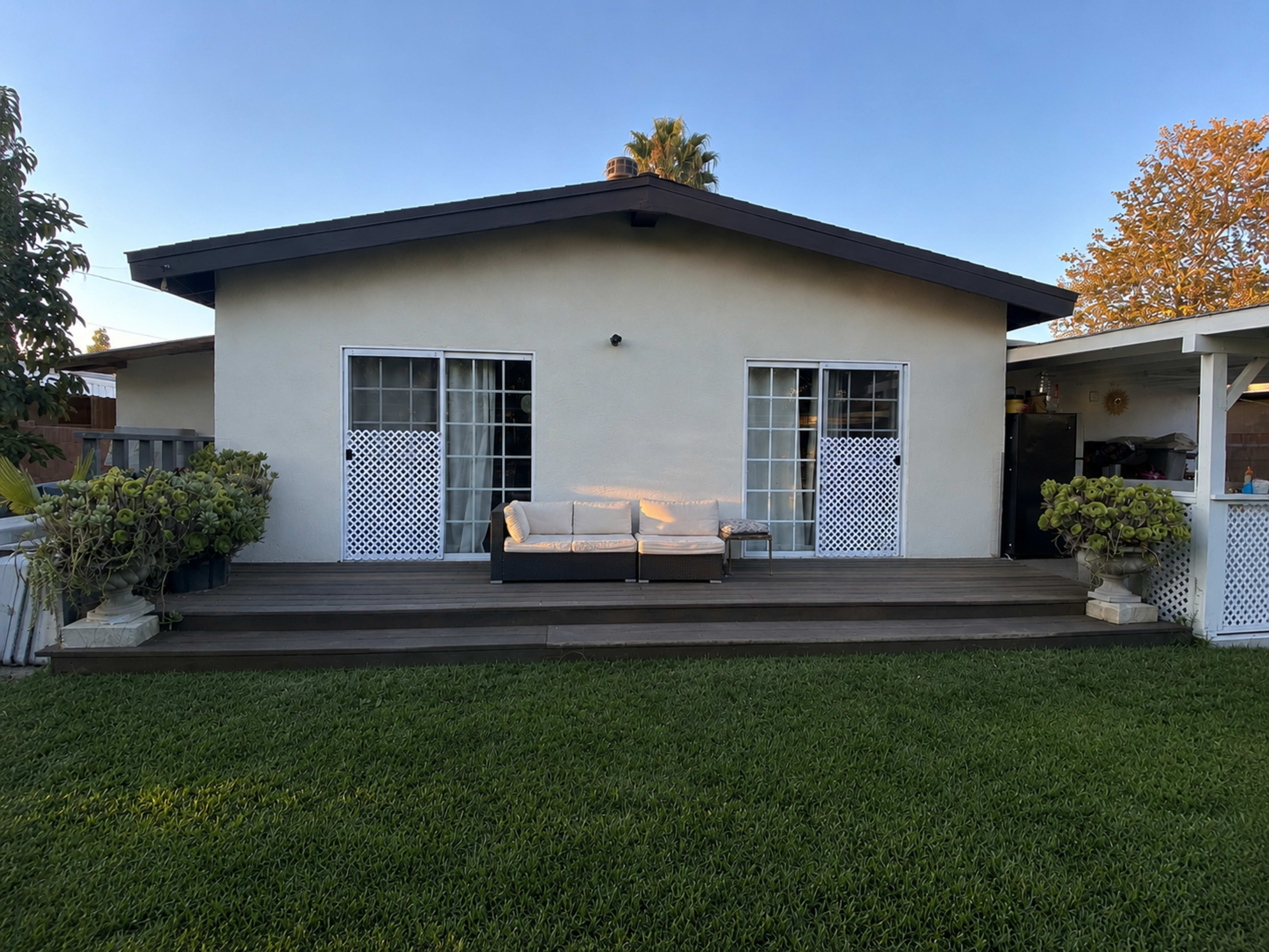 A single-story house features a wooden deck with a sofa and two glass doors leading inside, surrounded by well-maintained grass and a few potted plants.