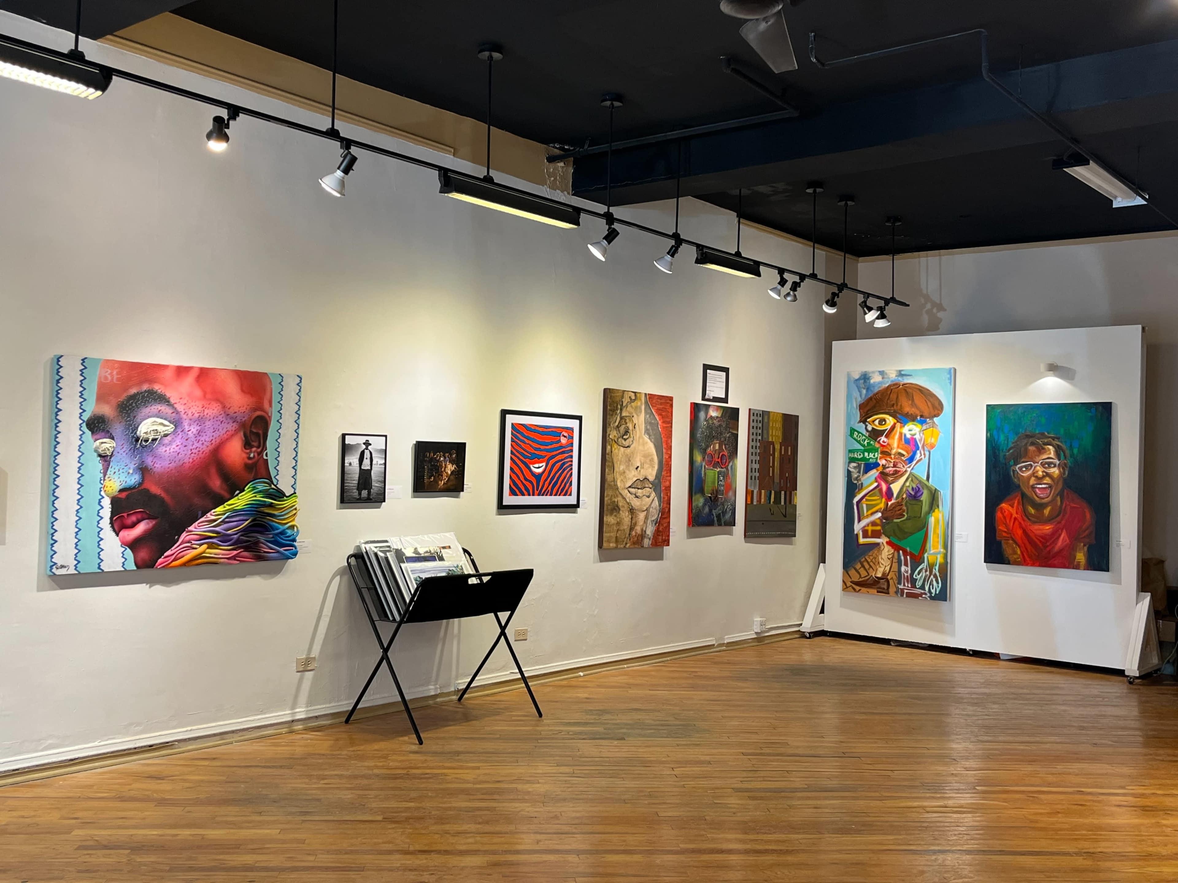 The image shows a gallery space with a variety of colorful artworks displayed on the walls and a black rack holding pamphlets or catalogs in the foreground.