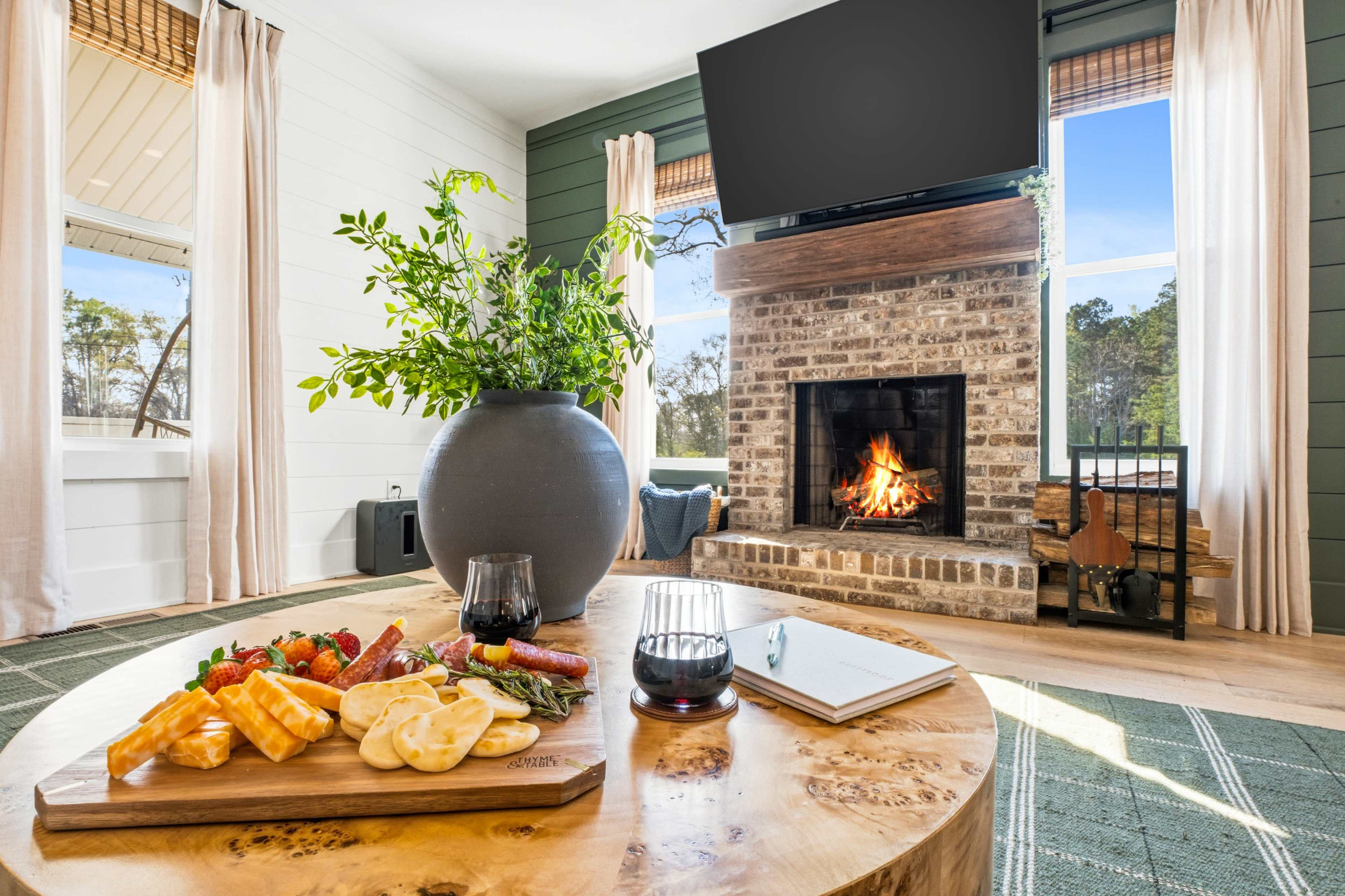 A cozy living room features a wooden coffee table with a charcuterie board, a large plant, and a fireplace with a lit fire, while a TV is mounted on the wall above.