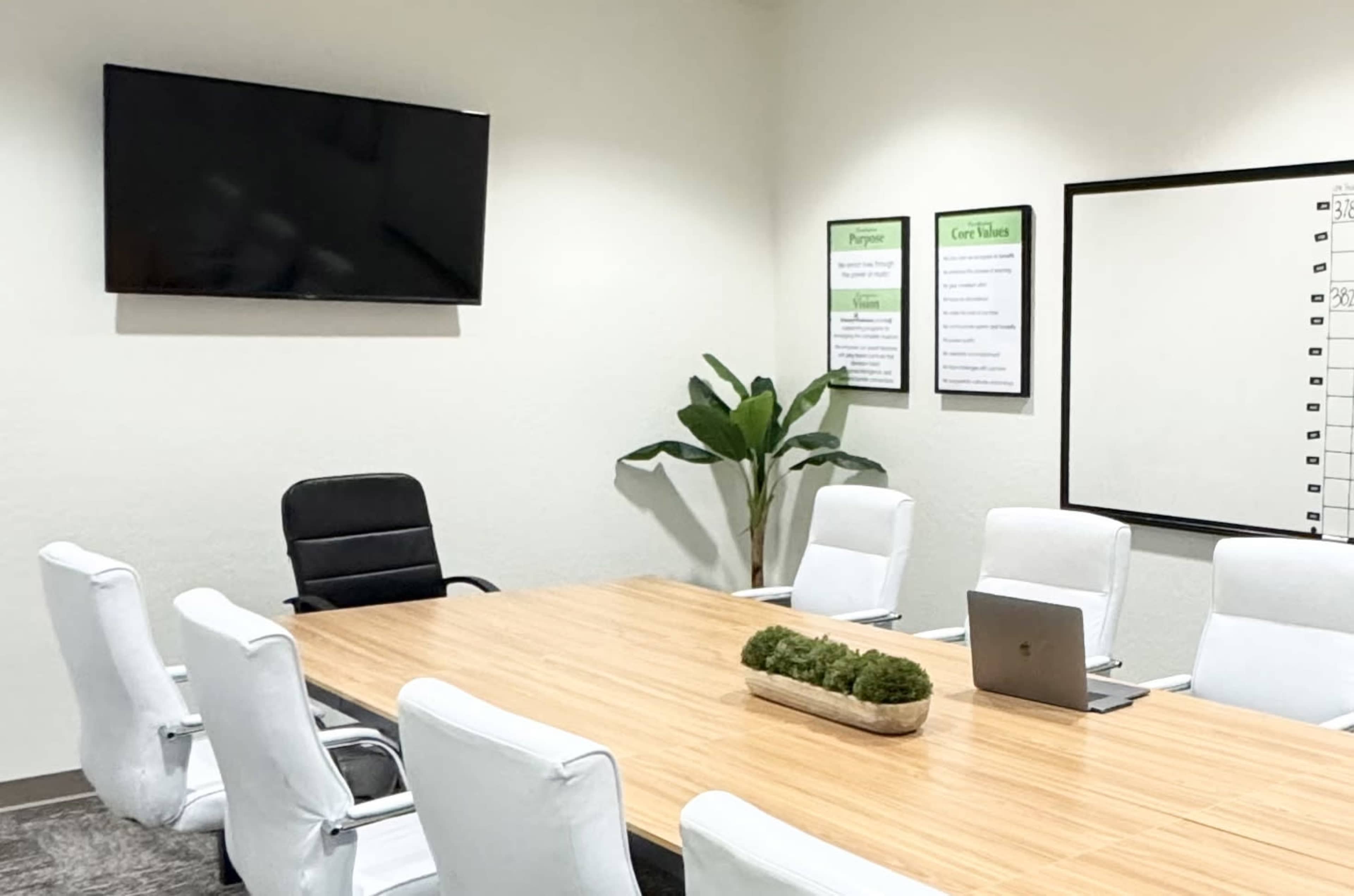 A modern conference room features a large wooden table surrounded by white chairs, a laptop on the table, and a wall-mounted TV.