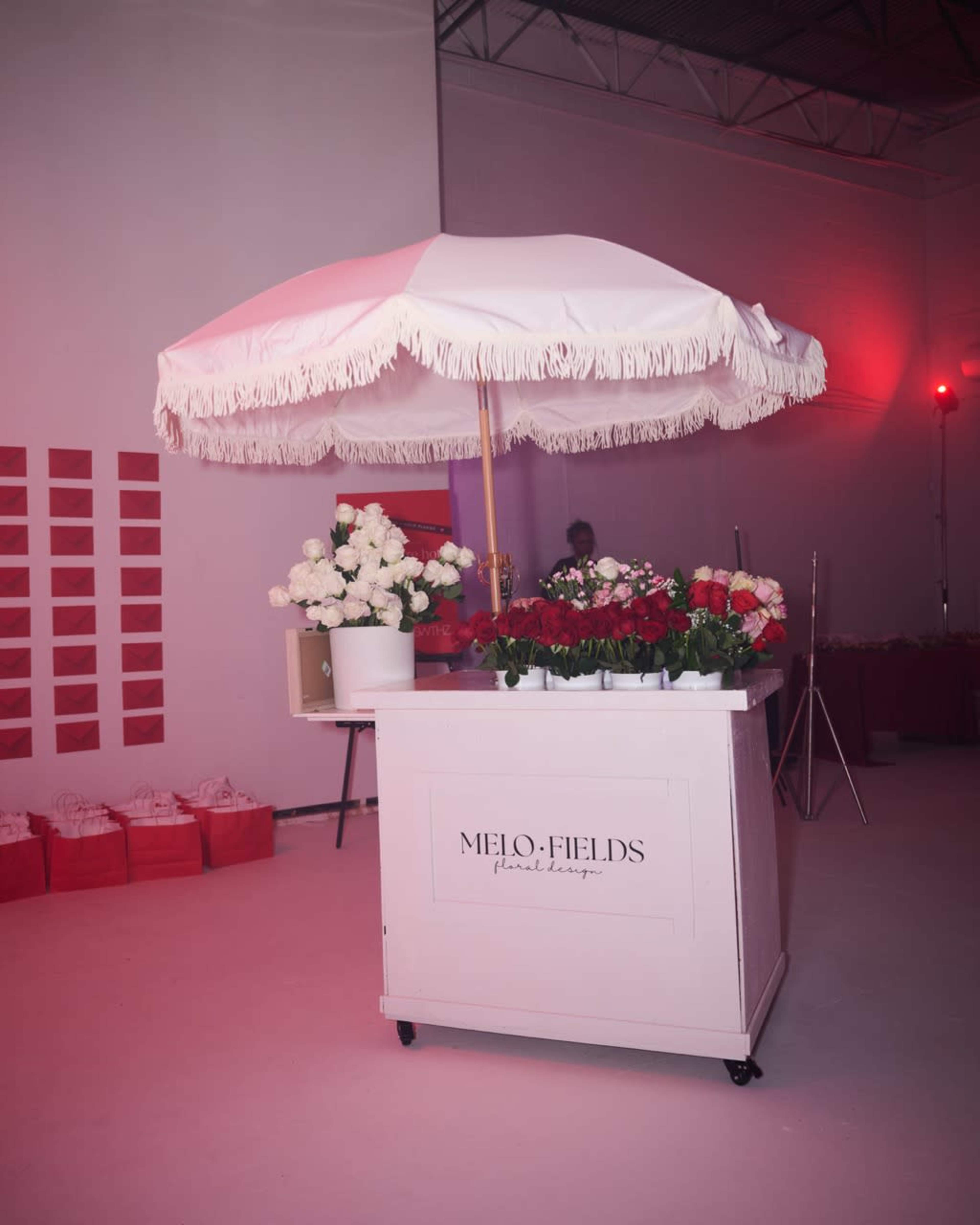 A white floral cart with an umbrella is filled with various flowers and positioned in a brightly lit venue.