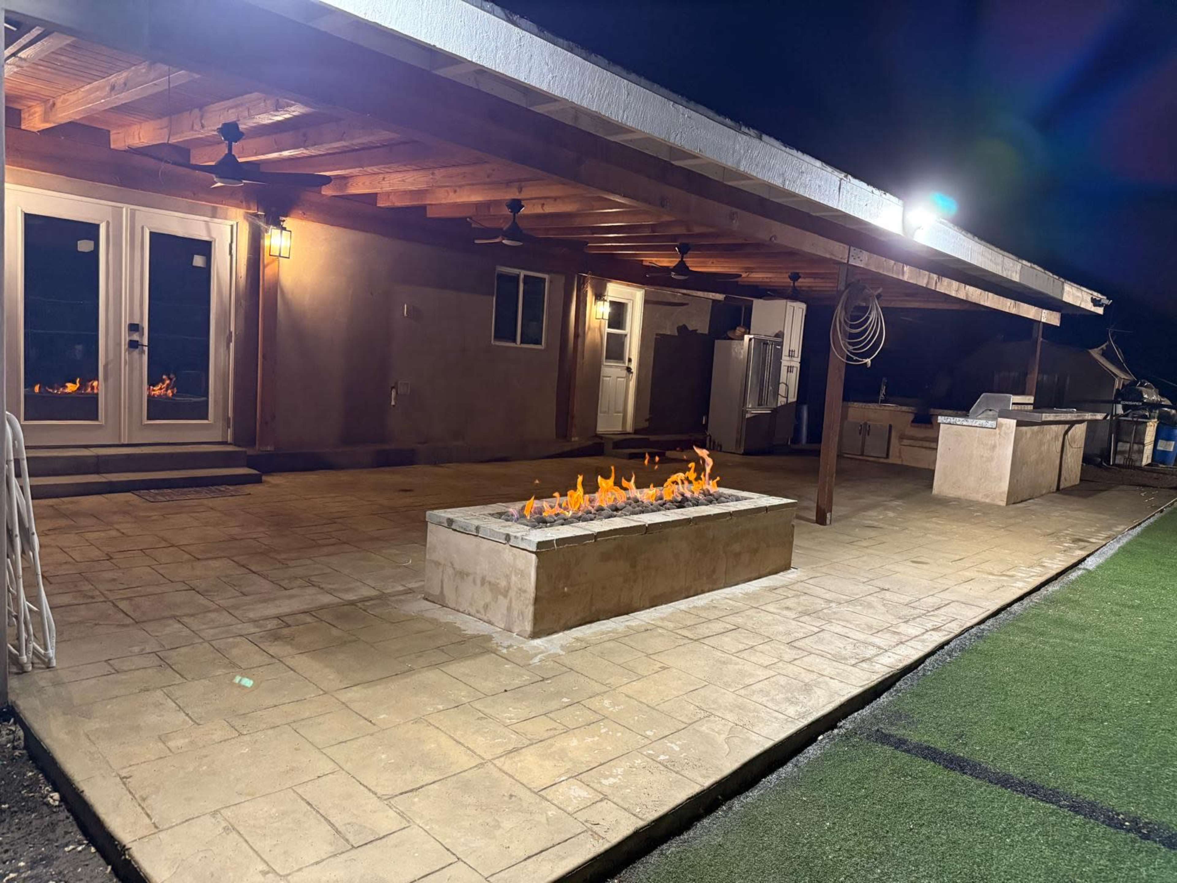 The image depicts a covered outdoor patio area at night, featuring a long fire pit with flames, adjacent to a kitchen space equipped with a grill and counter.