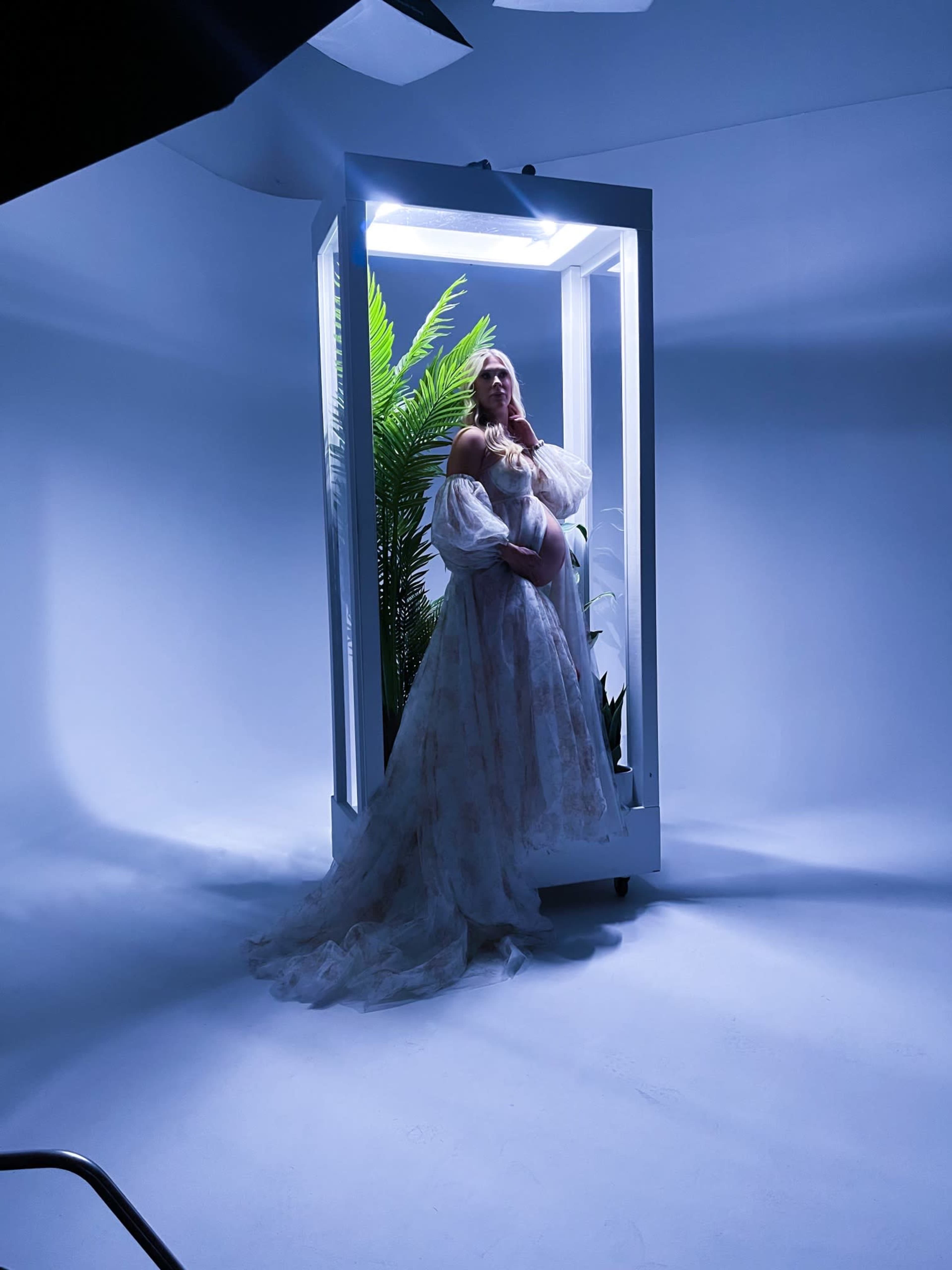 A woman in a flowing gown stands inside a white frame with plants, illuminated by soft light against a blue backdrop.