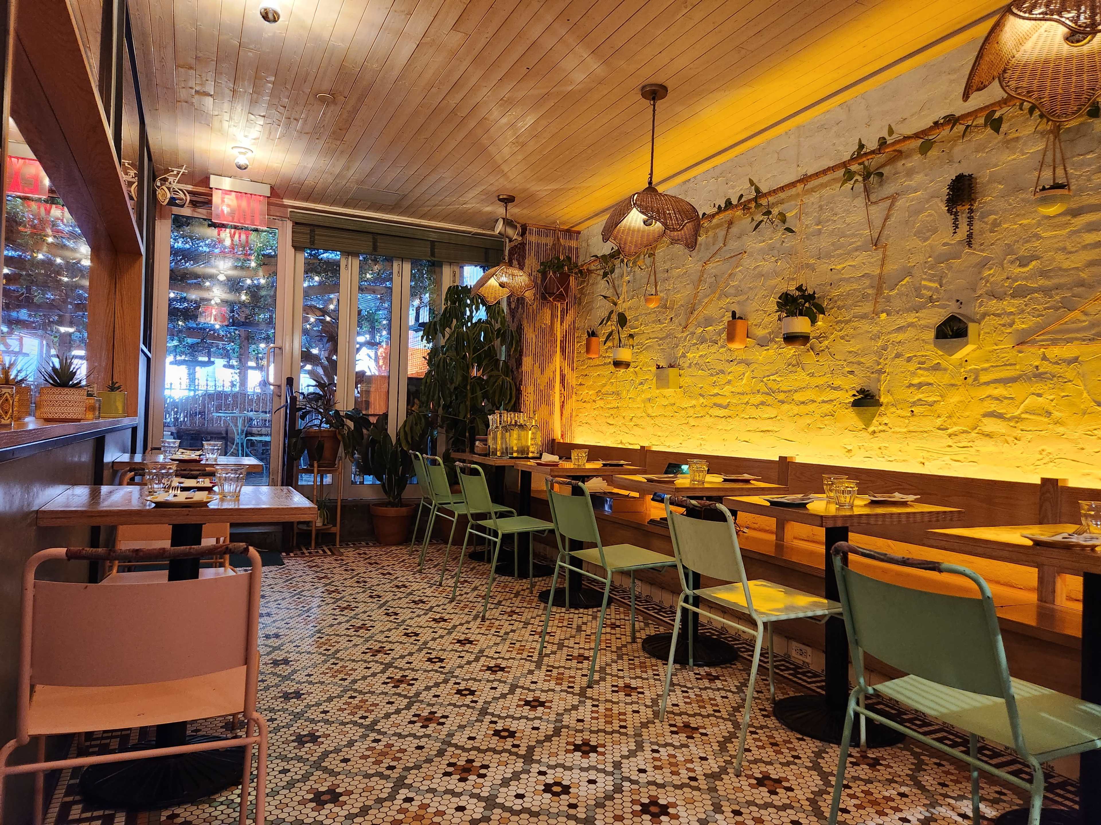 A brightly lit restaurant interior featuring a tiled floor, wooden tables, and plants along the walls.