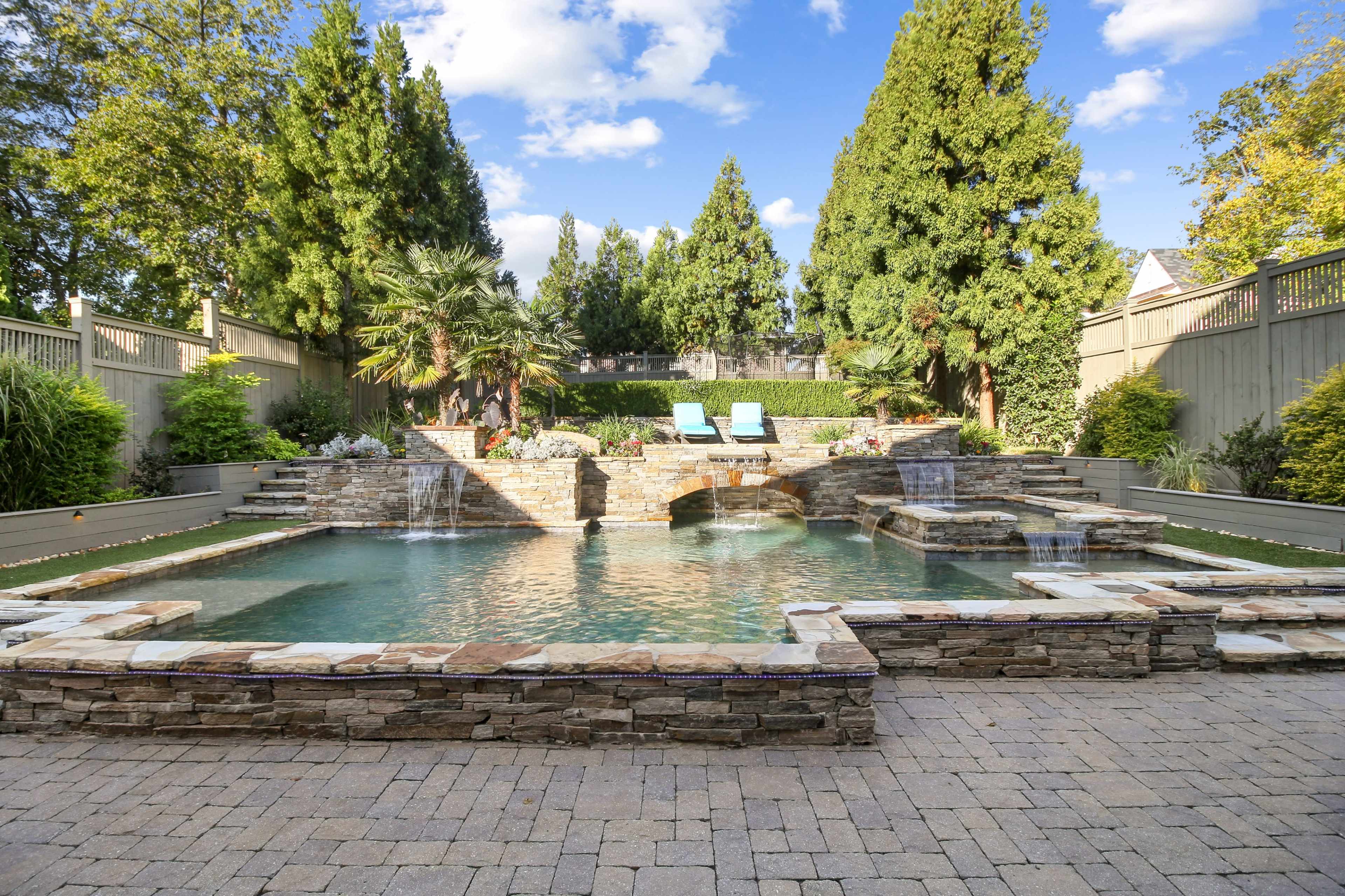 A landscaped backyard featuring a stone-accented swimming pool with waterfalls, surrounded by lush greenery and lounge chairs.