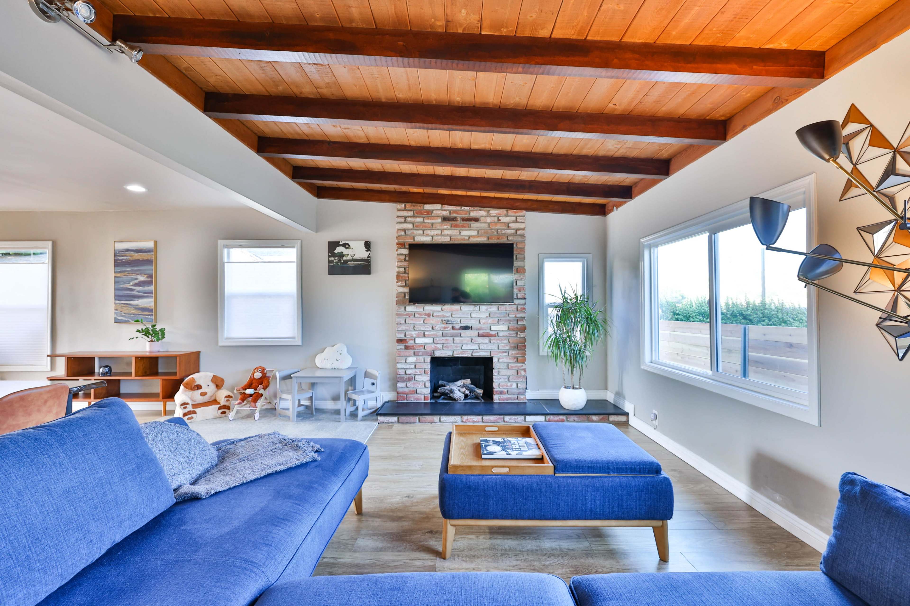 A modern living room featuring exposed wooden beams, a brick fireplace, a large TV, and blue upholstered furniture.