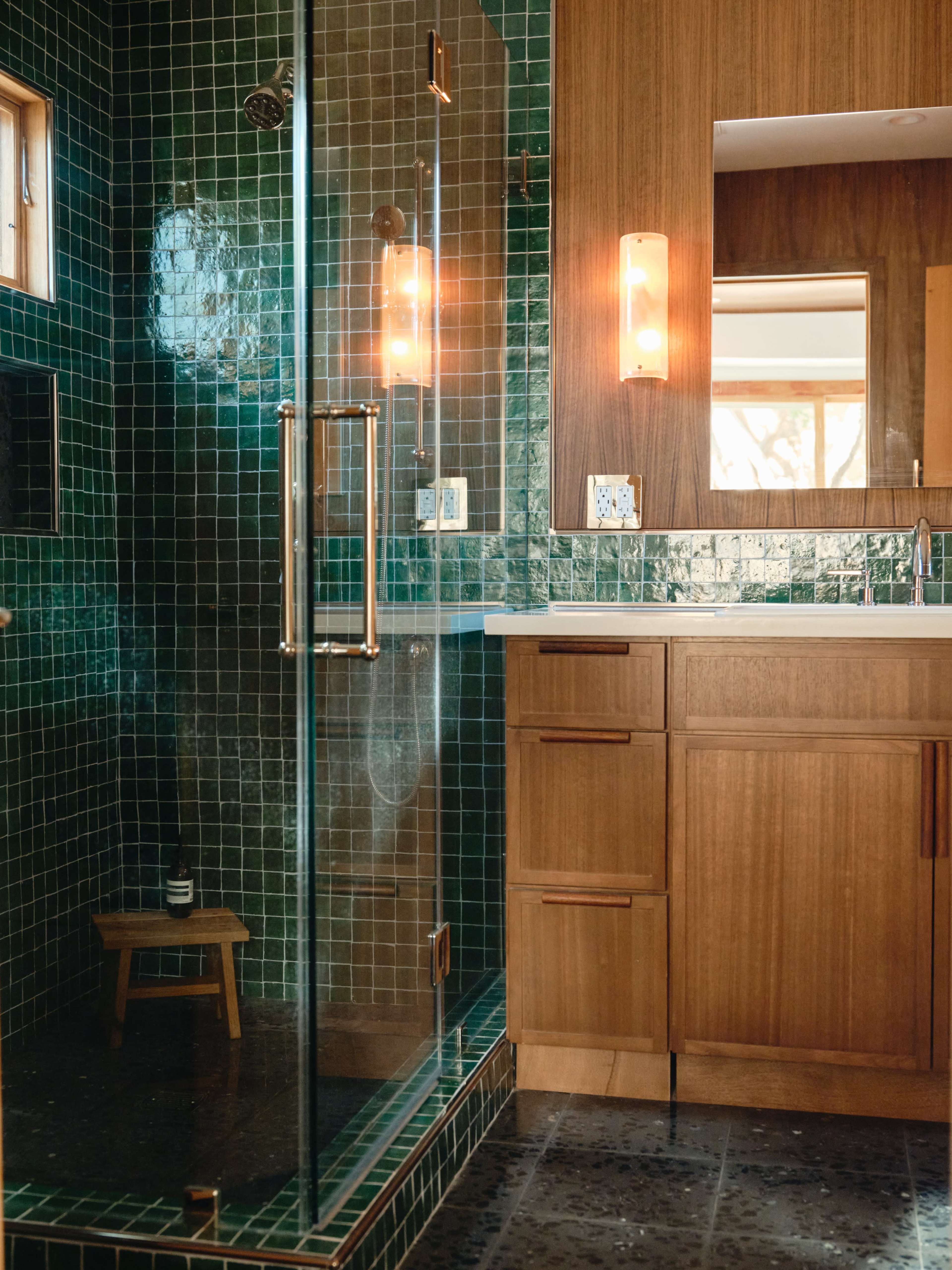 The image shows a modern bathroom featuring a glass shower enclosure, green tiled walls, wooden cabinetry, and a well-lit mirror area.