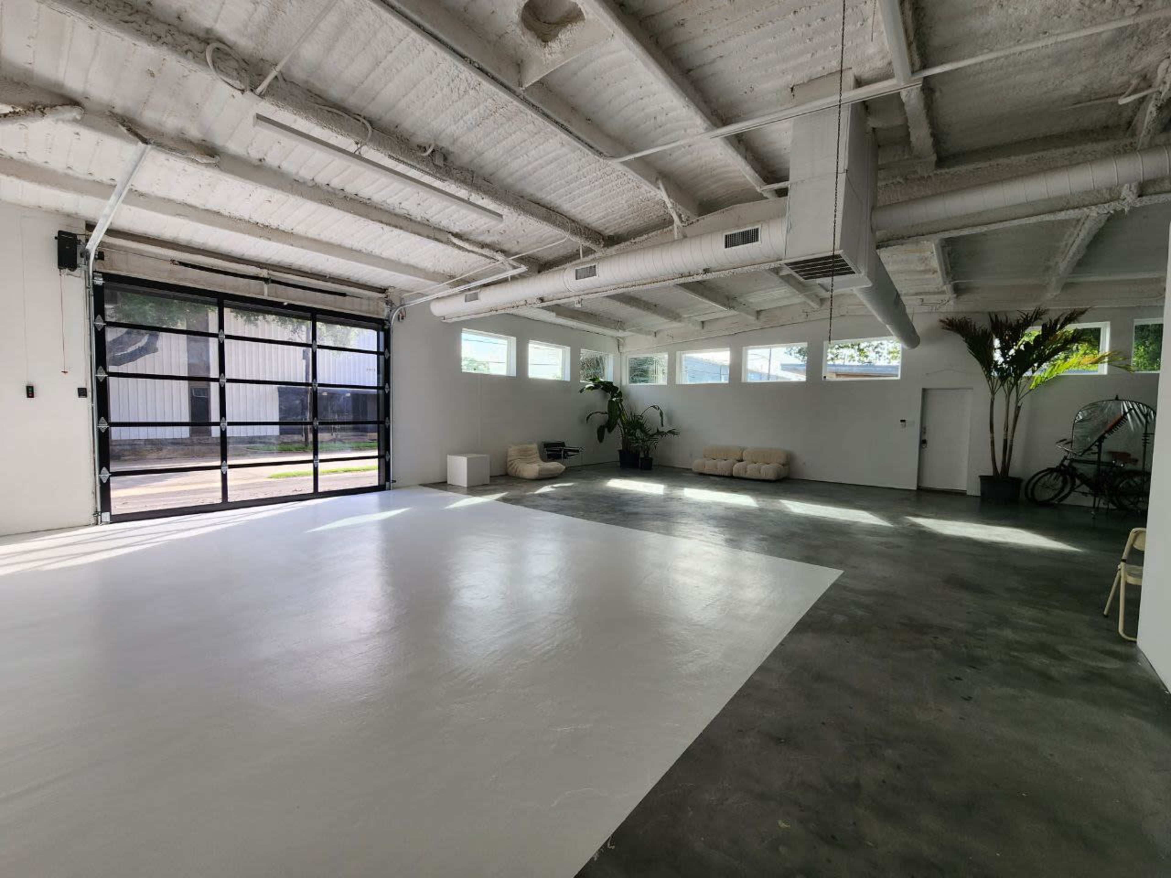 The image depicts a spacious garage with a polished white floor, large glass doors, and minimal furnishings including a sofa and potted plants.