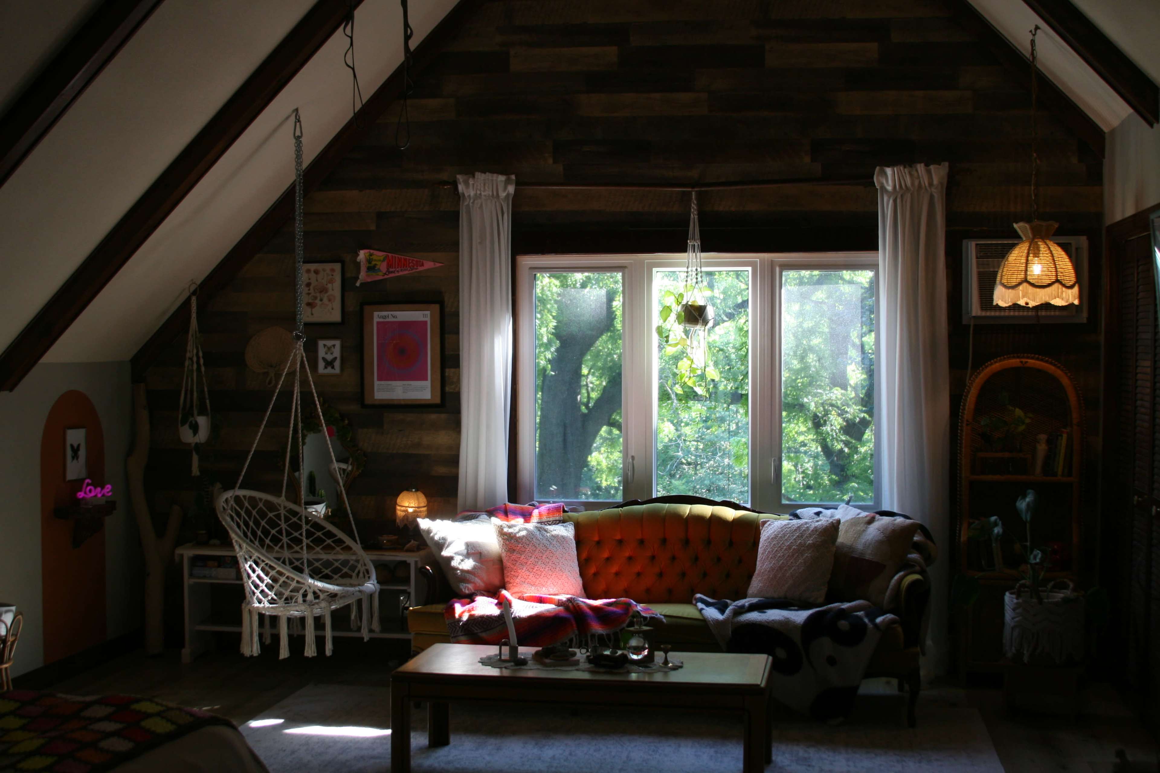 The image shows a cozy living room with a hanging chair, a bright orange couch, and large windows that let in natural light, surrounded by trees.
