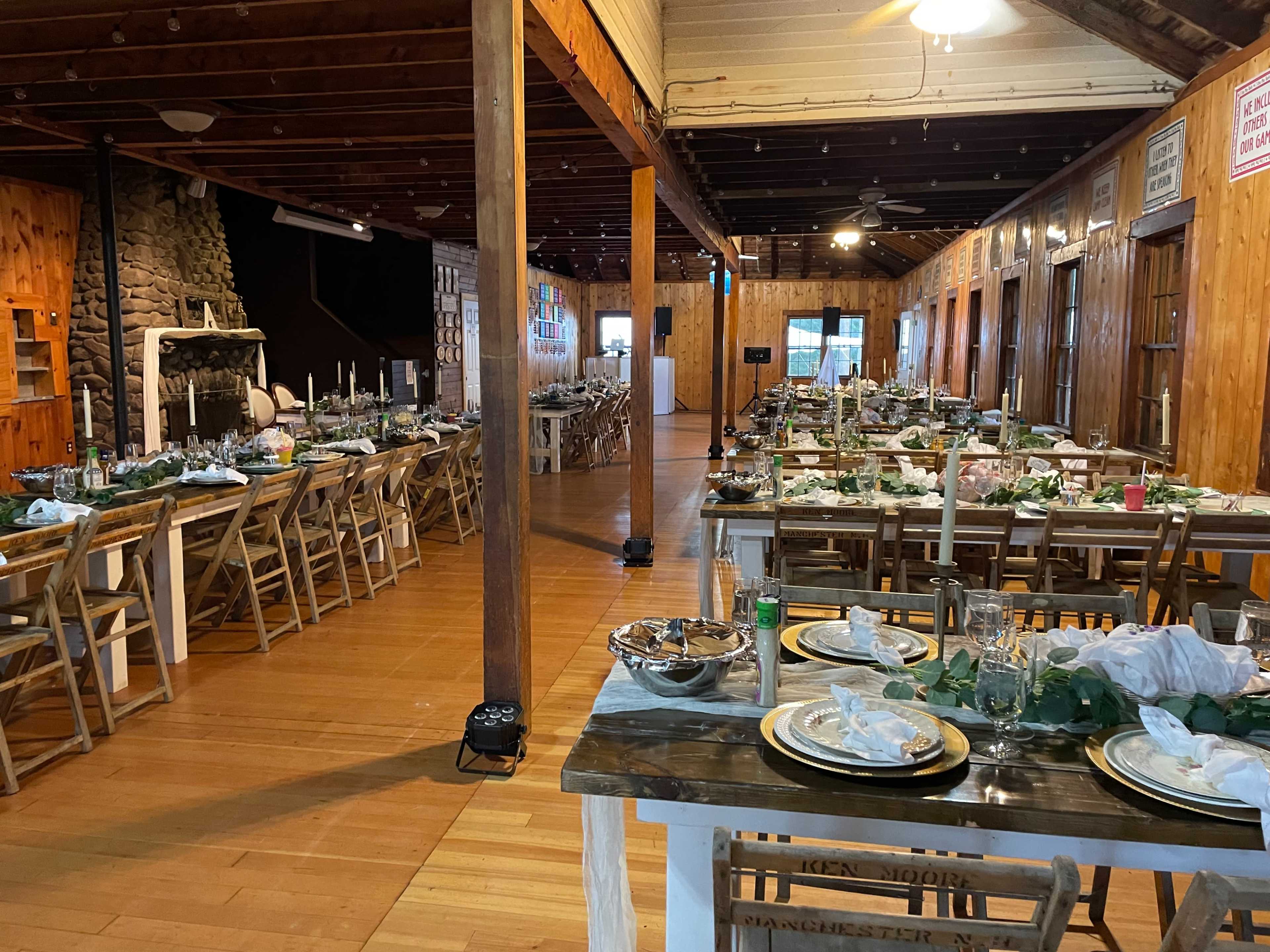 A spacious dining hall with several long tables set for a meal, featuring plates, glasses, and decorations, under wooden beams and a stone fireplace.