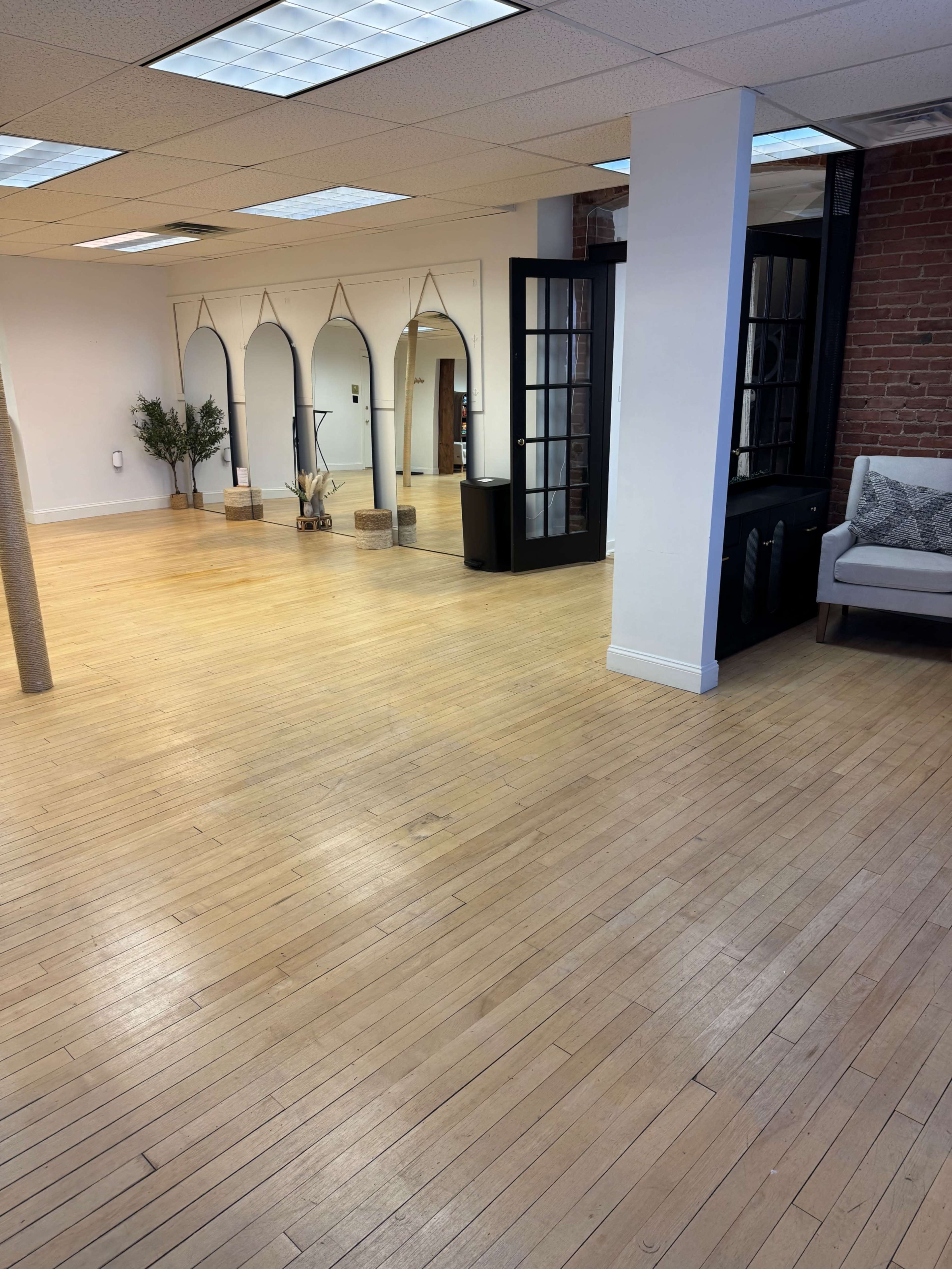 The image shows a spacious, well-lit room with wooden flooring, a large mirror with an arch design, and a small seating area with a couch.