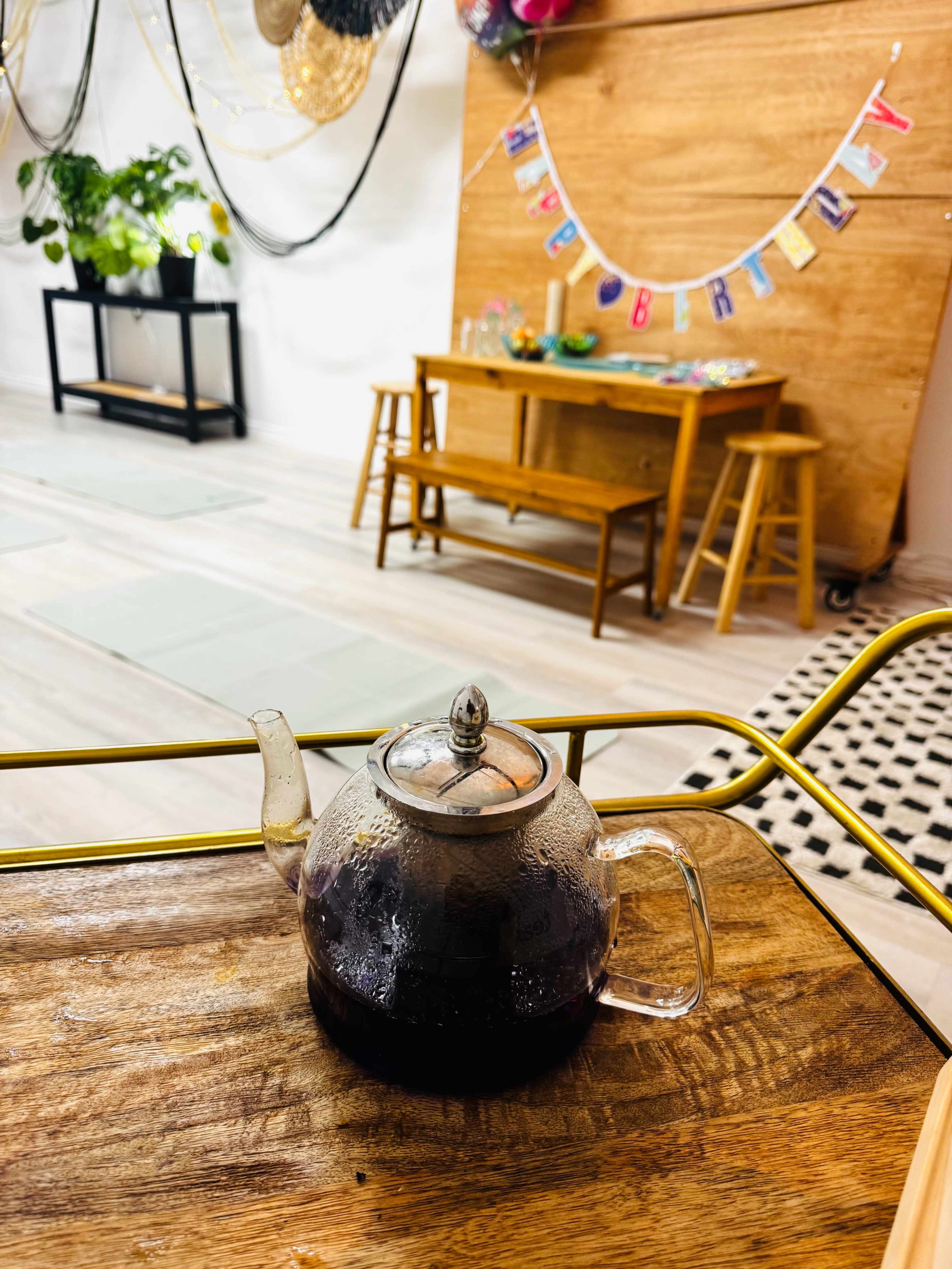 A glass teapot filled with dark tea sits on a wooden table in a room decorated for a birthday celebration, featuring a festive banner and various seating arrangements.