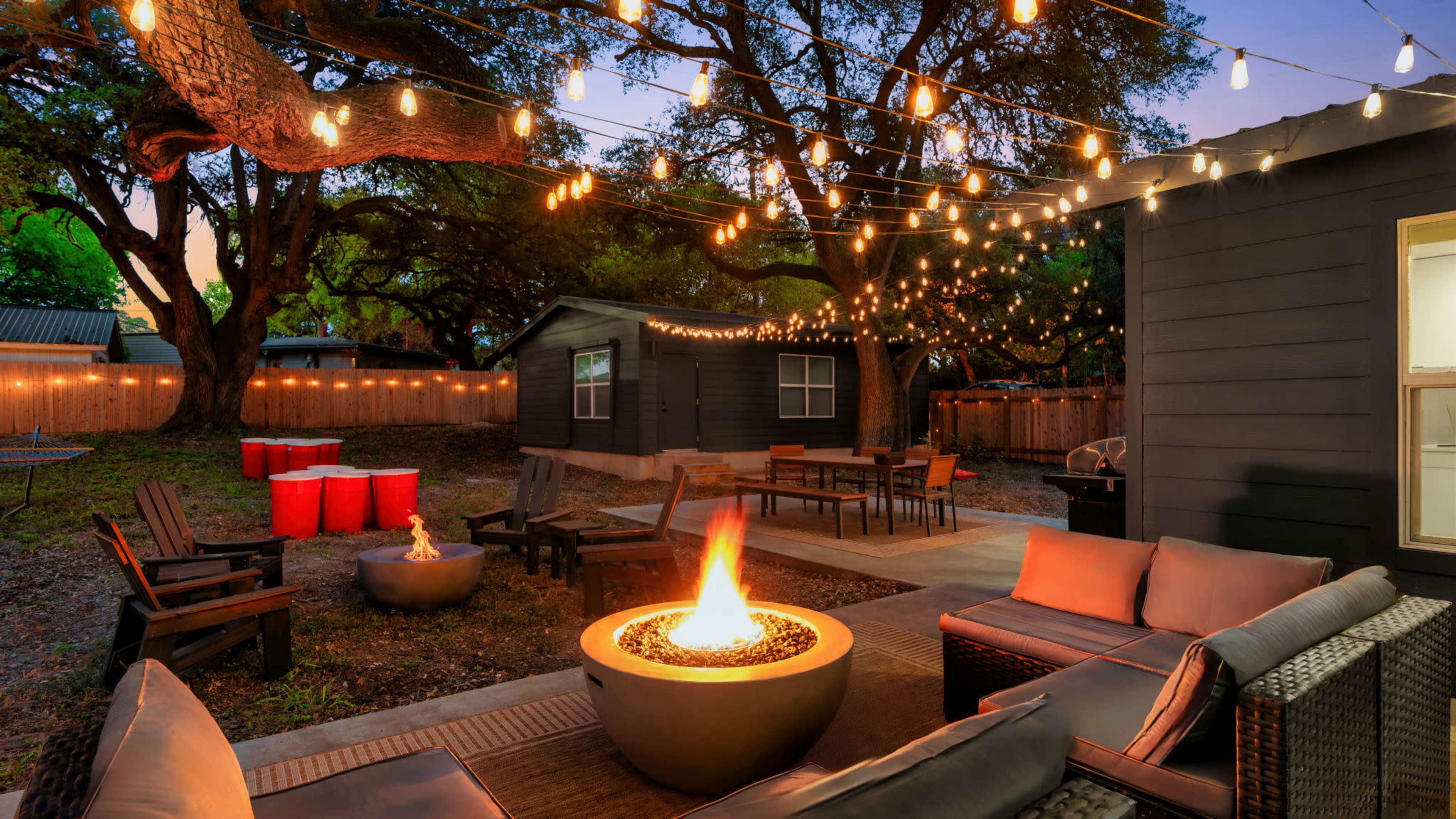A cozy outdoor patio features a fire pit surrounded by seating, string lights overhead, and colorful barrels in the background.