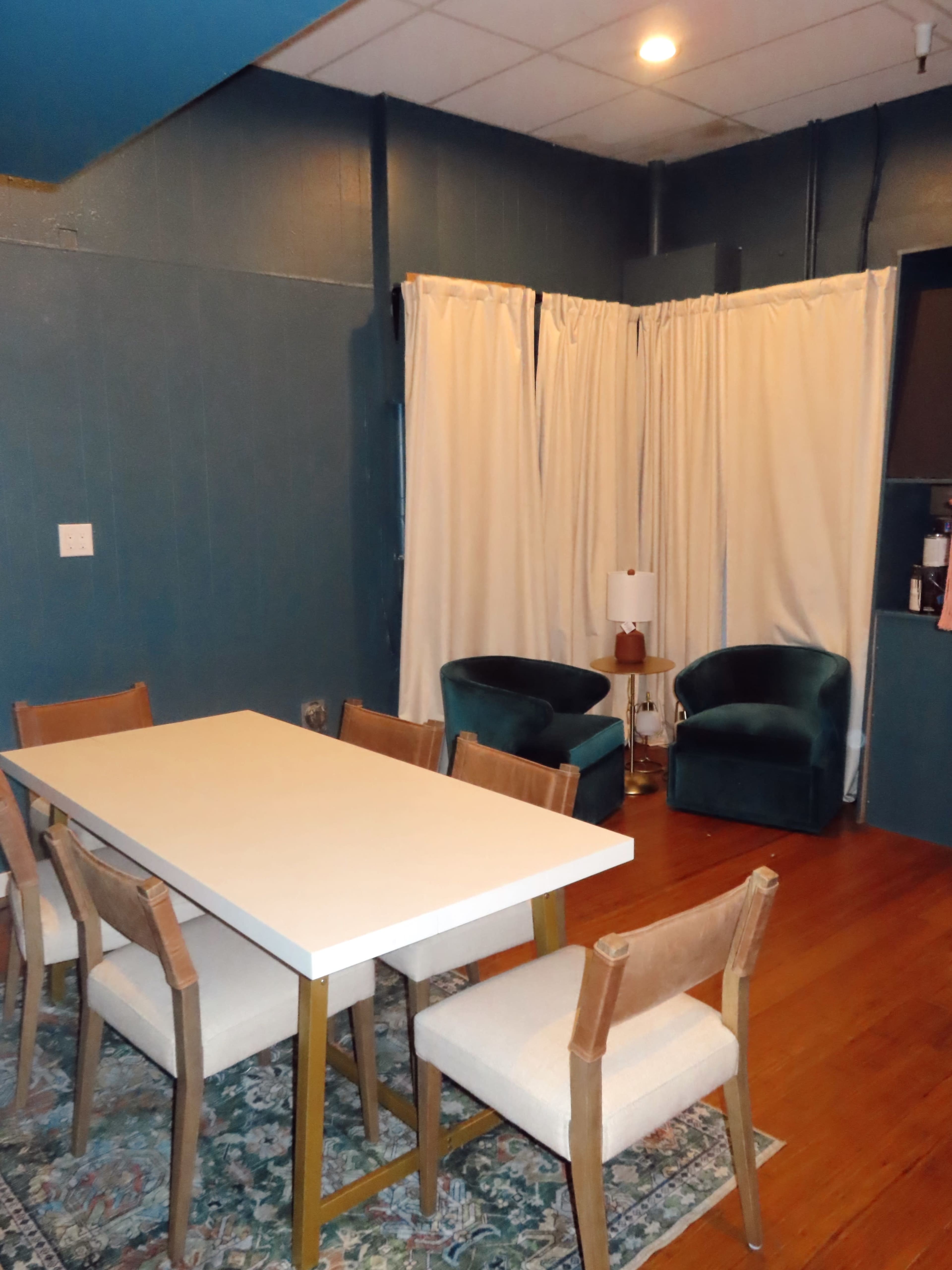 A dining area features a white table surrounded by wooden chairs, with two teal armchairs and a lamp in a corner behind curtains.