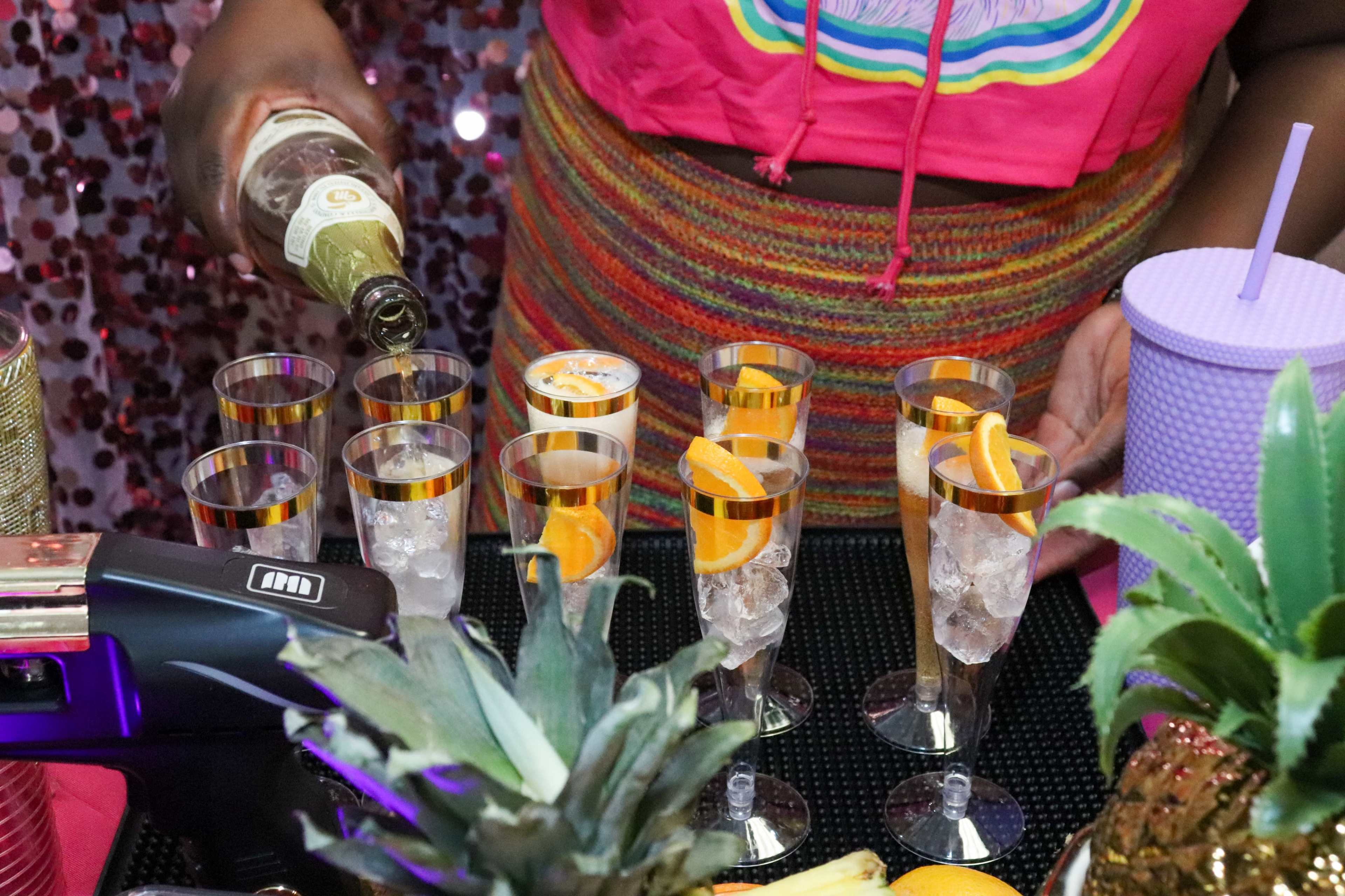 A person pours a sparkling beverage into a row of decorated glasses filled with ice and garnished with slices of orange.
