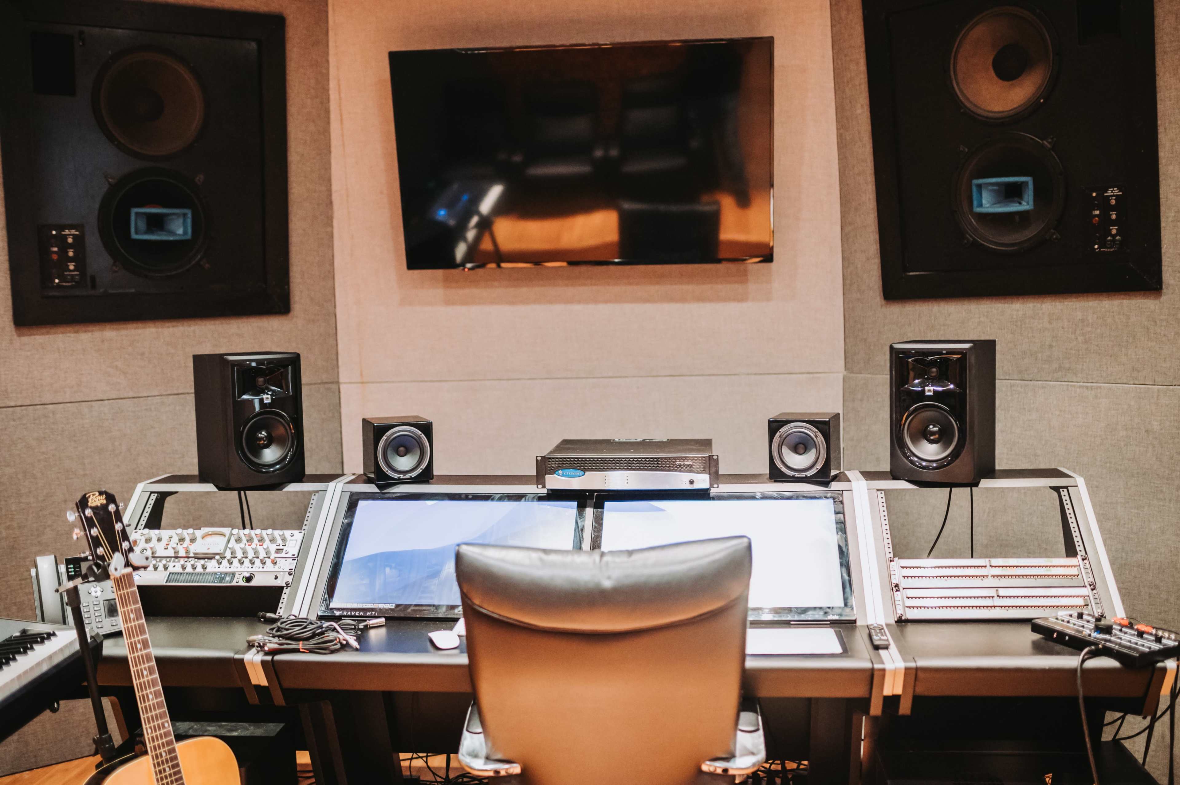 A music production studio features a desk with two computer monitors, audio equipment, and speakers, surrounded by soundproof walls.