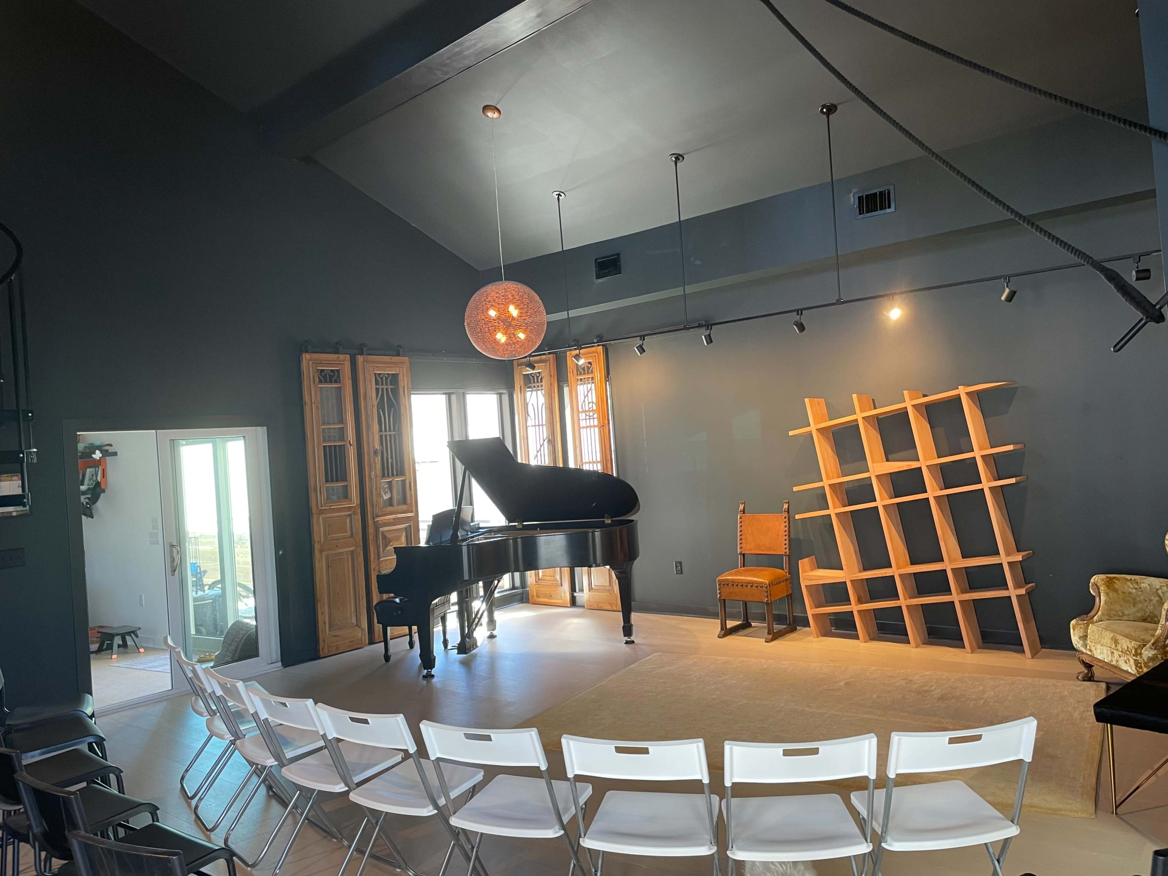 A black piano is positioned in the center of a room with gray walls, accompanied by a wooden chair and a shelving unit against the wall, while white folding chairs are set up in front.