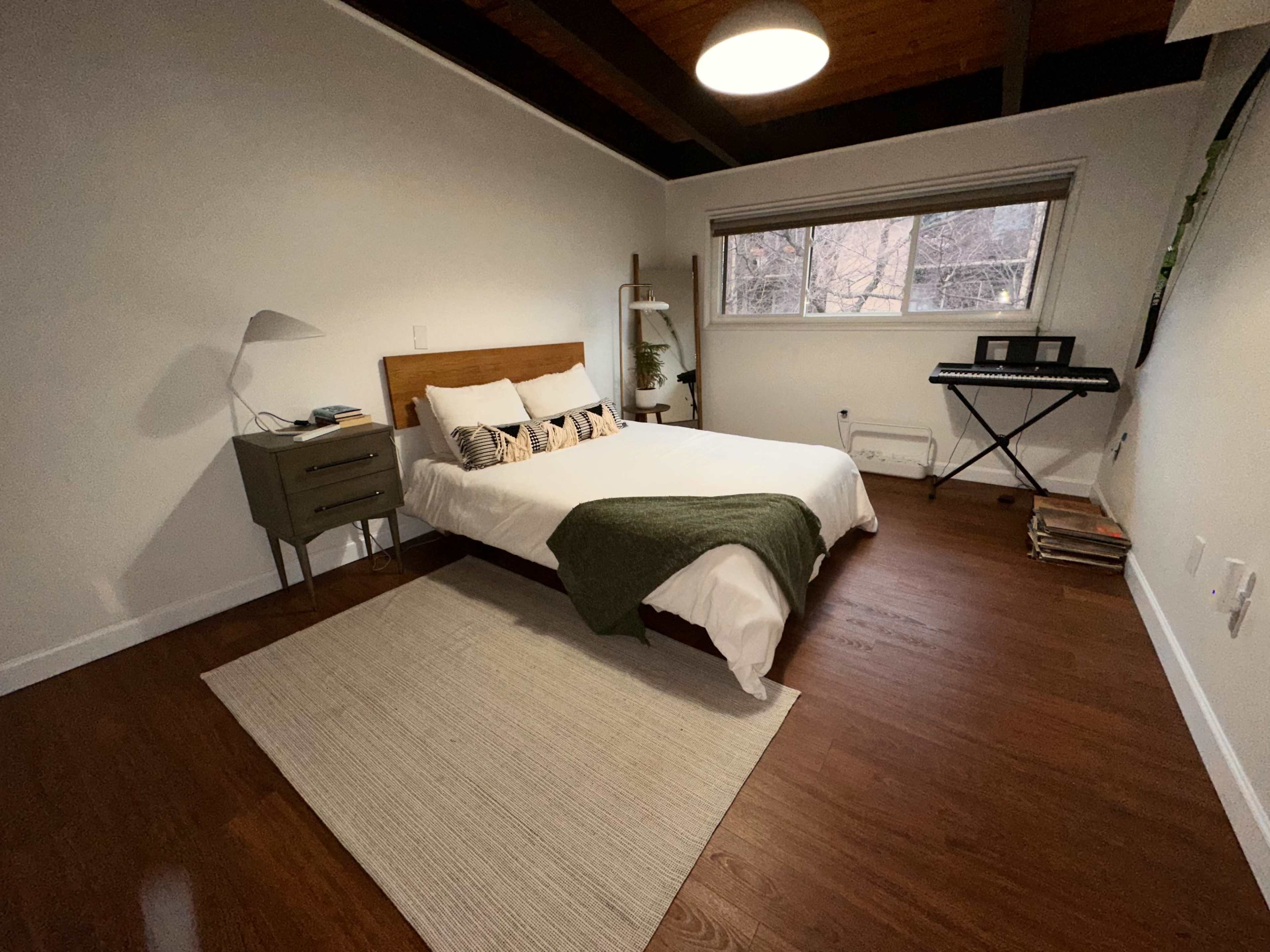 A cozy bedroom featuring a bed with a green throw, a bedside table with a lamp, and a keyboard against the wall, all set on hardwood floors with a large window.
