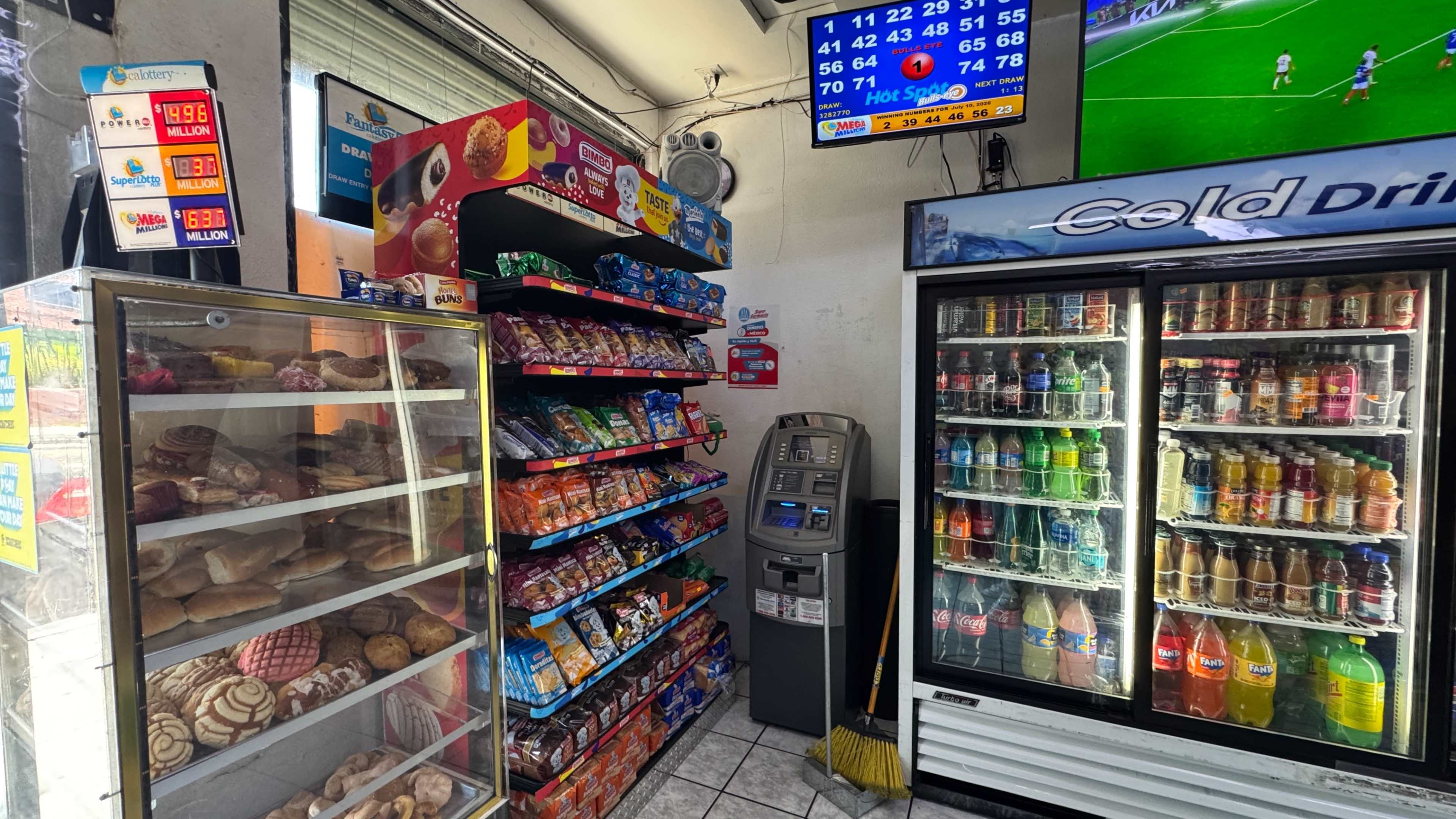 The image shows a convenience store interior with shelves stocked with snacks, a glass display case filled with pastries, an ATM, and a vending refrigerator filled with various beverages, alongside a screen displaying sports scores.
