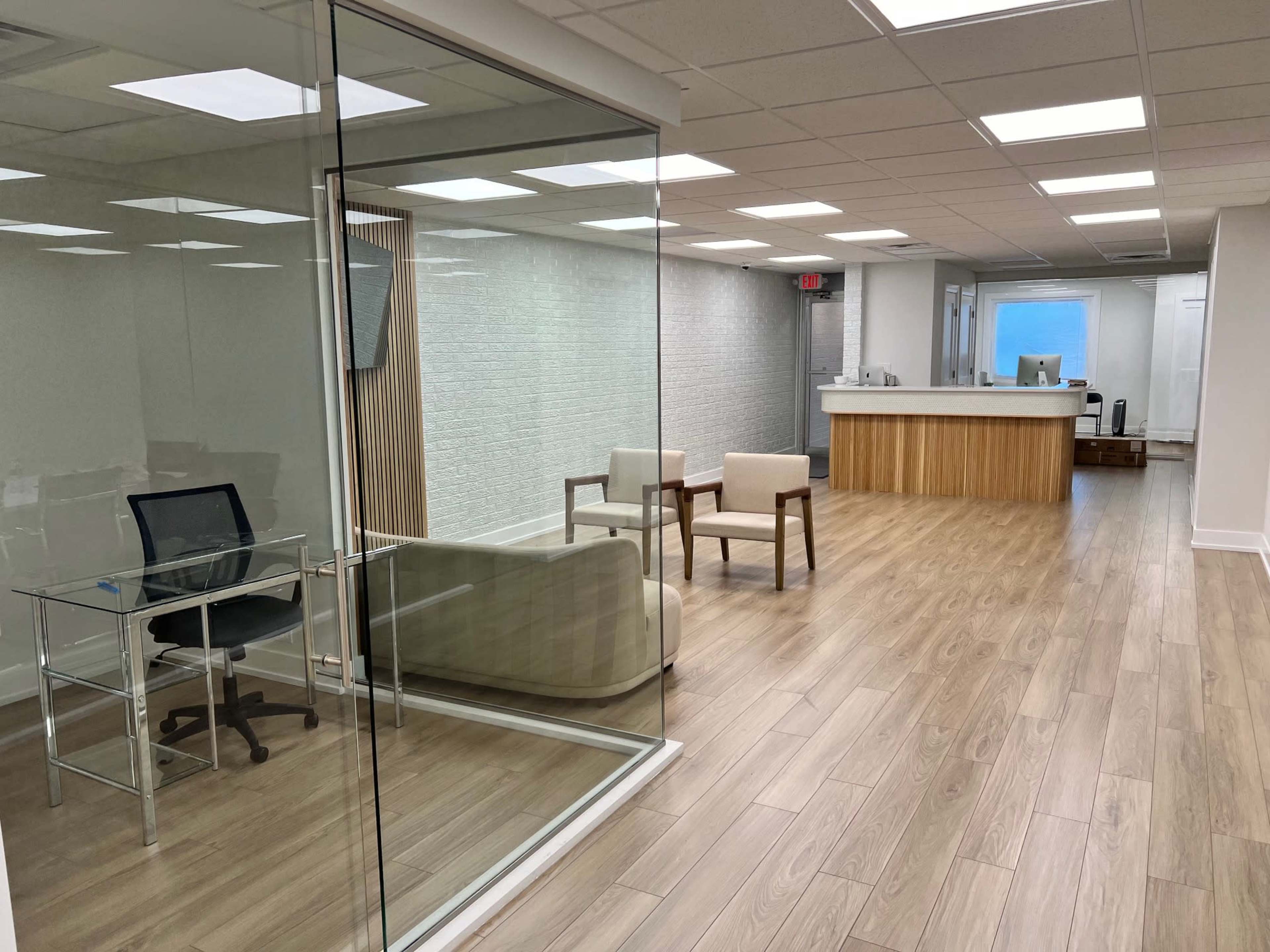 A modern office interior featuring a glass-walled meeting room, two chairs, a desk, and a reception area with a wooden counter.