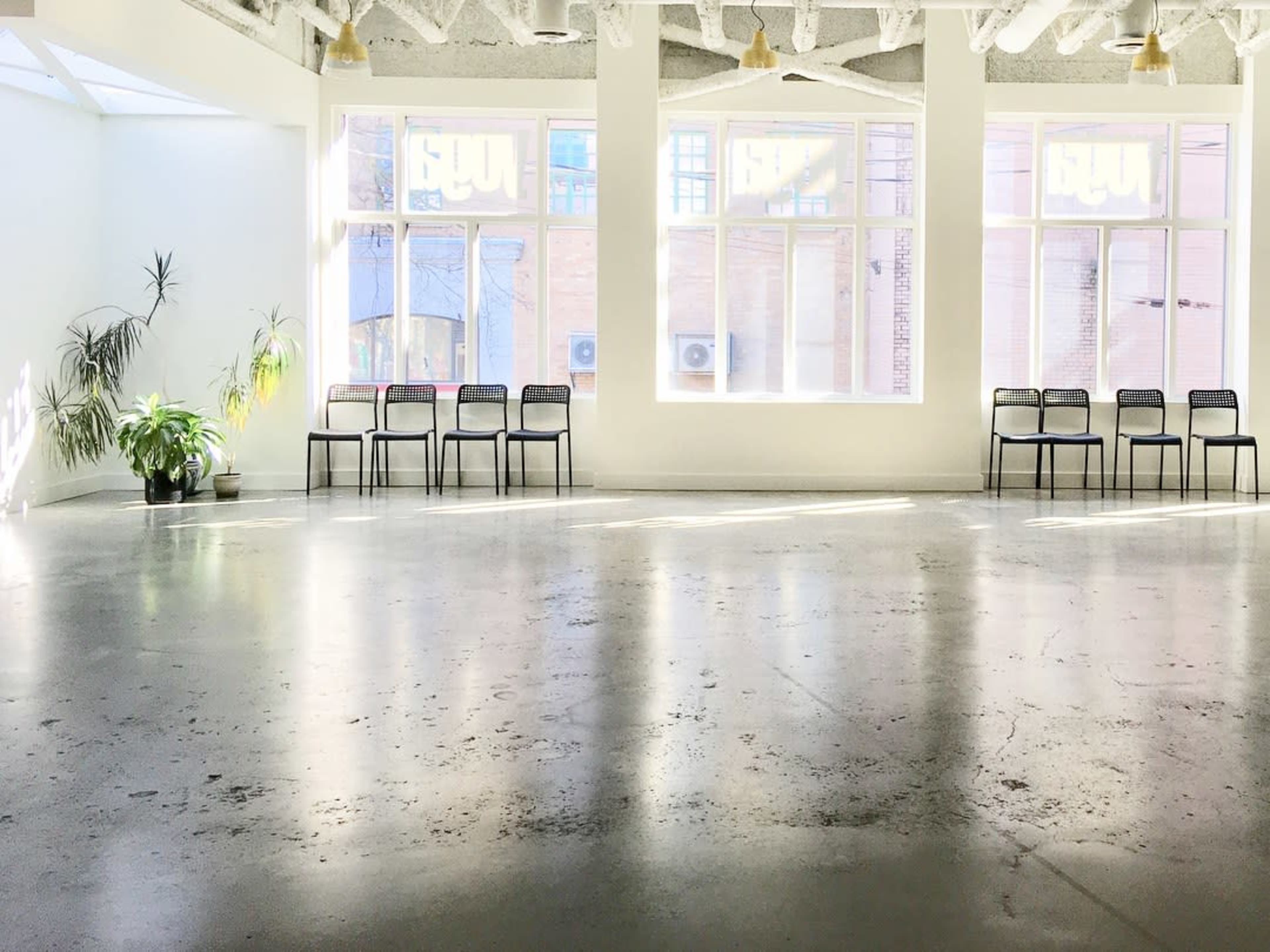 An empty room with concrete floors, several large windows allowing natural light, and a row of black chairs along one wall.