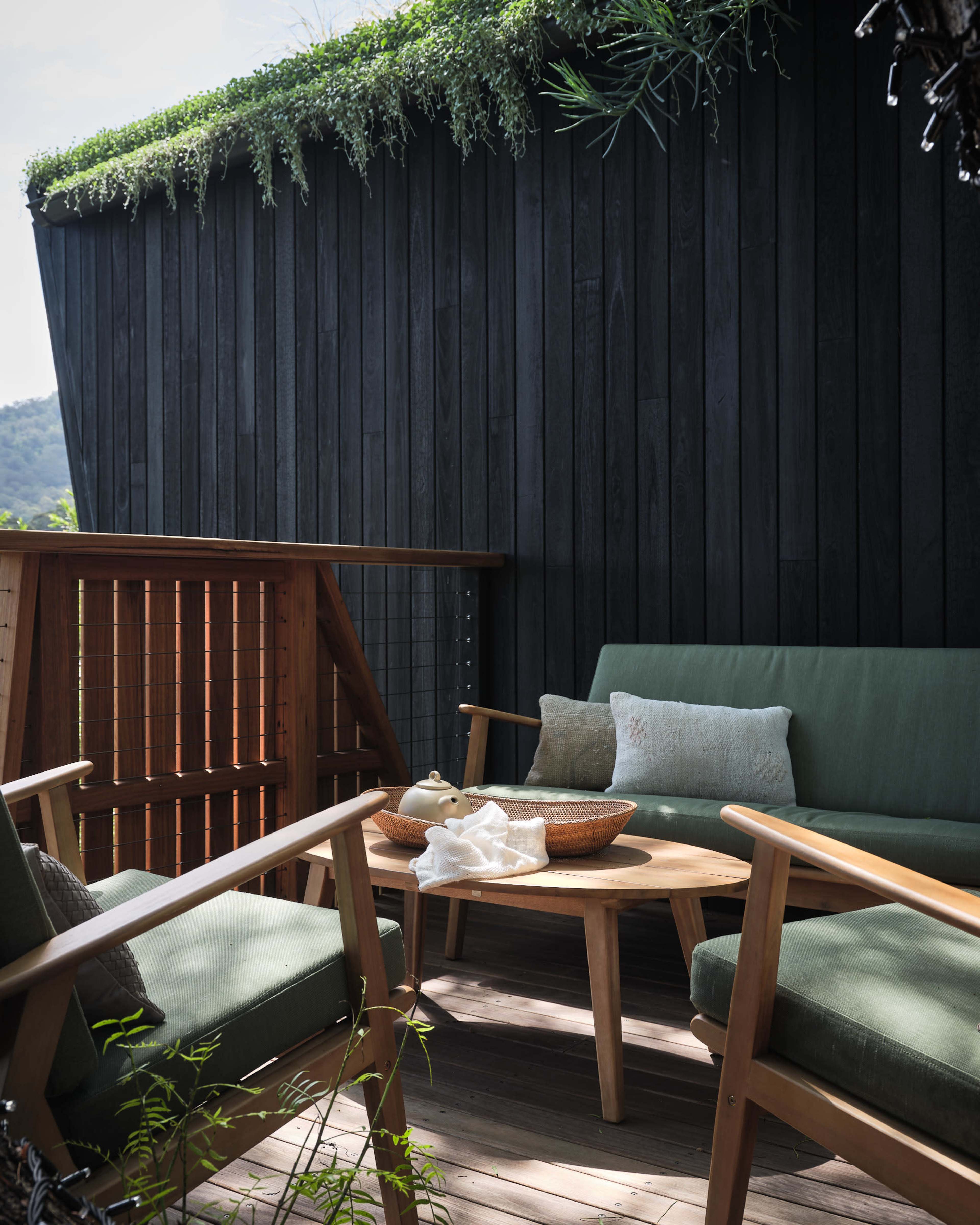 The image depicts a small outdoor seating area with green cushions on wooden chairs surrounding a circular table, complemented by a vertical garden on a dark wooden wall.