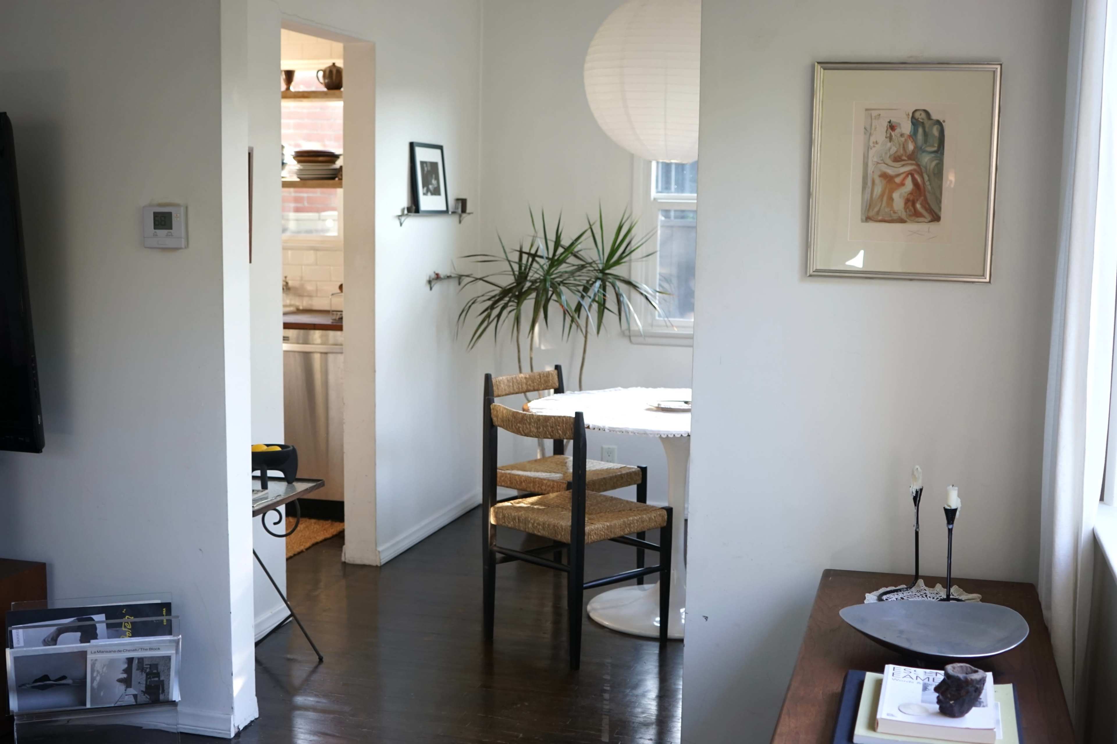 A bright, minimalistic living space featuring a dining area with a round table and two chairs, a doorway leading to a kitchen, and a small decorative table.
