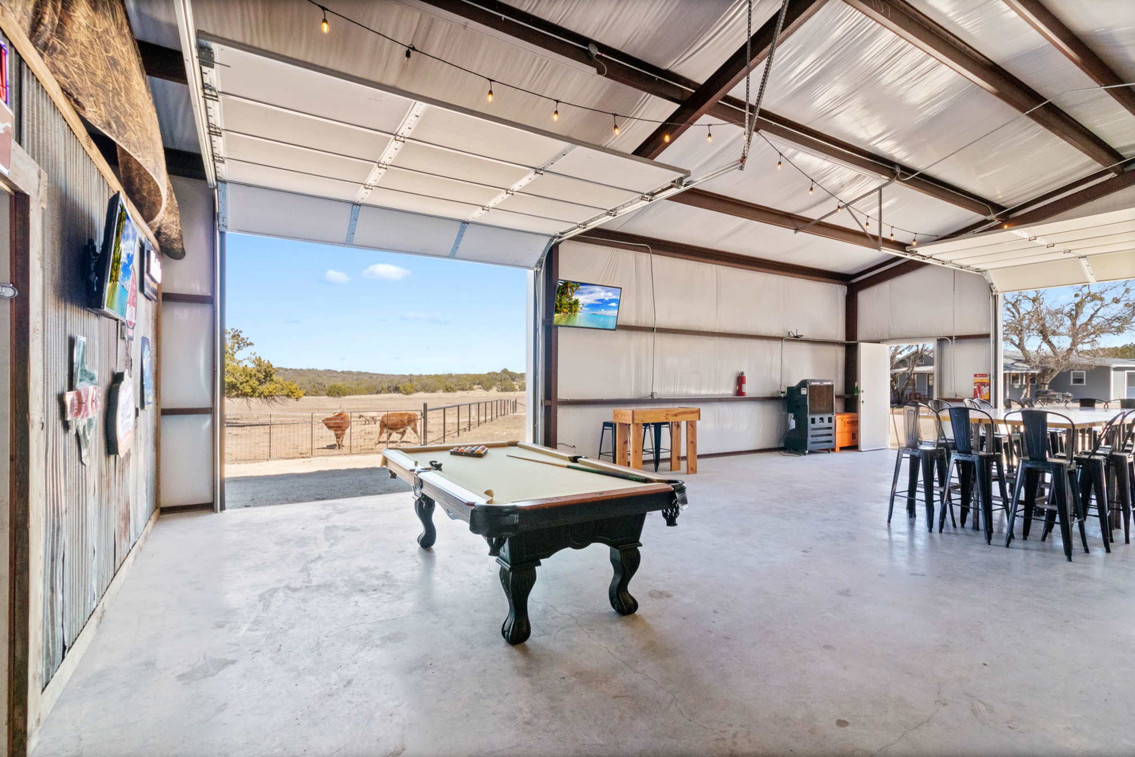 A spacious indoor area features a pool table, bar seating, and large open doors leading to a scenic outdoor view.