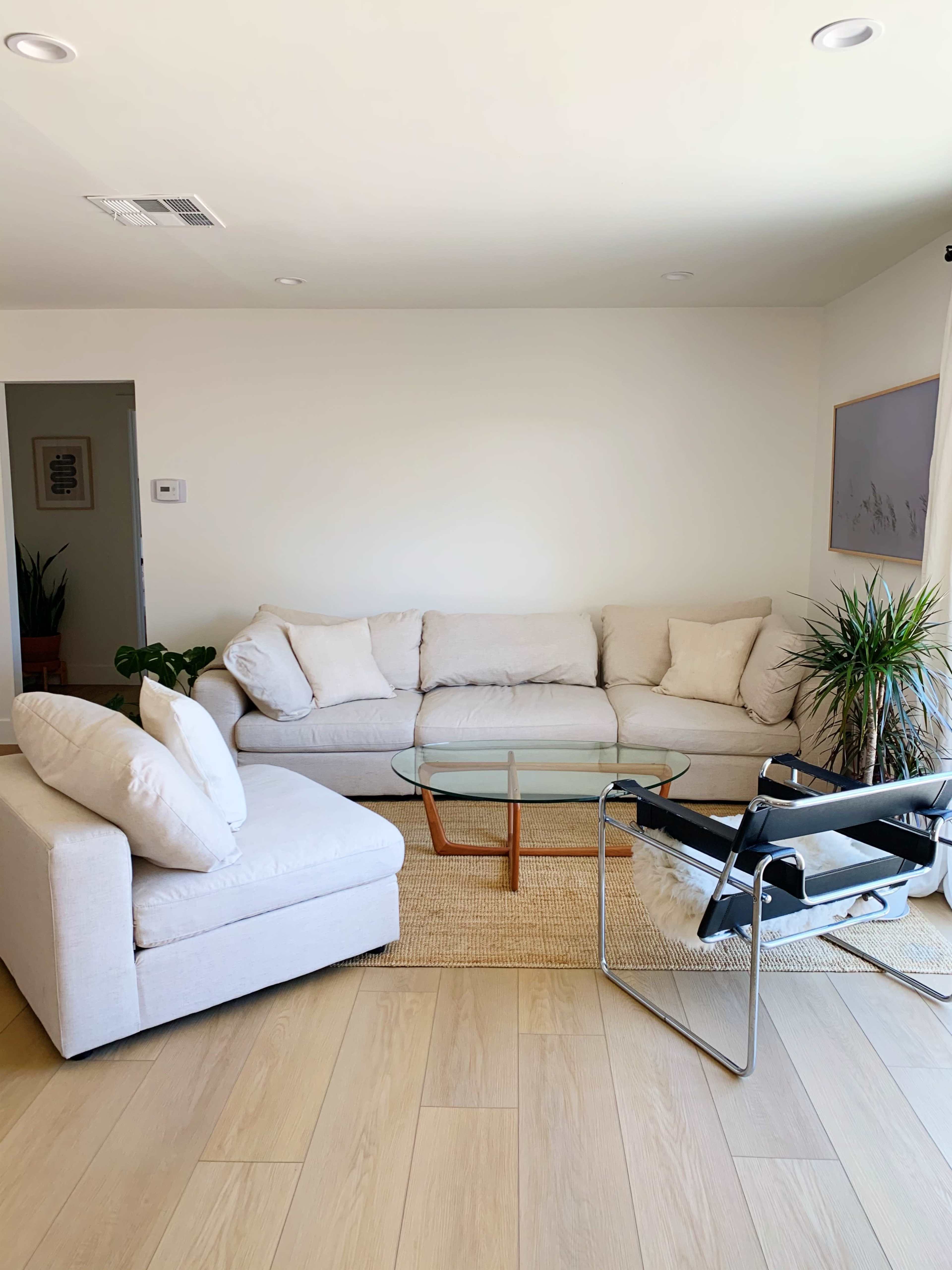 A light-colored living room features a sectional couch, a glass coffee table, and a black chair with a cushion, all set on a woven rug.