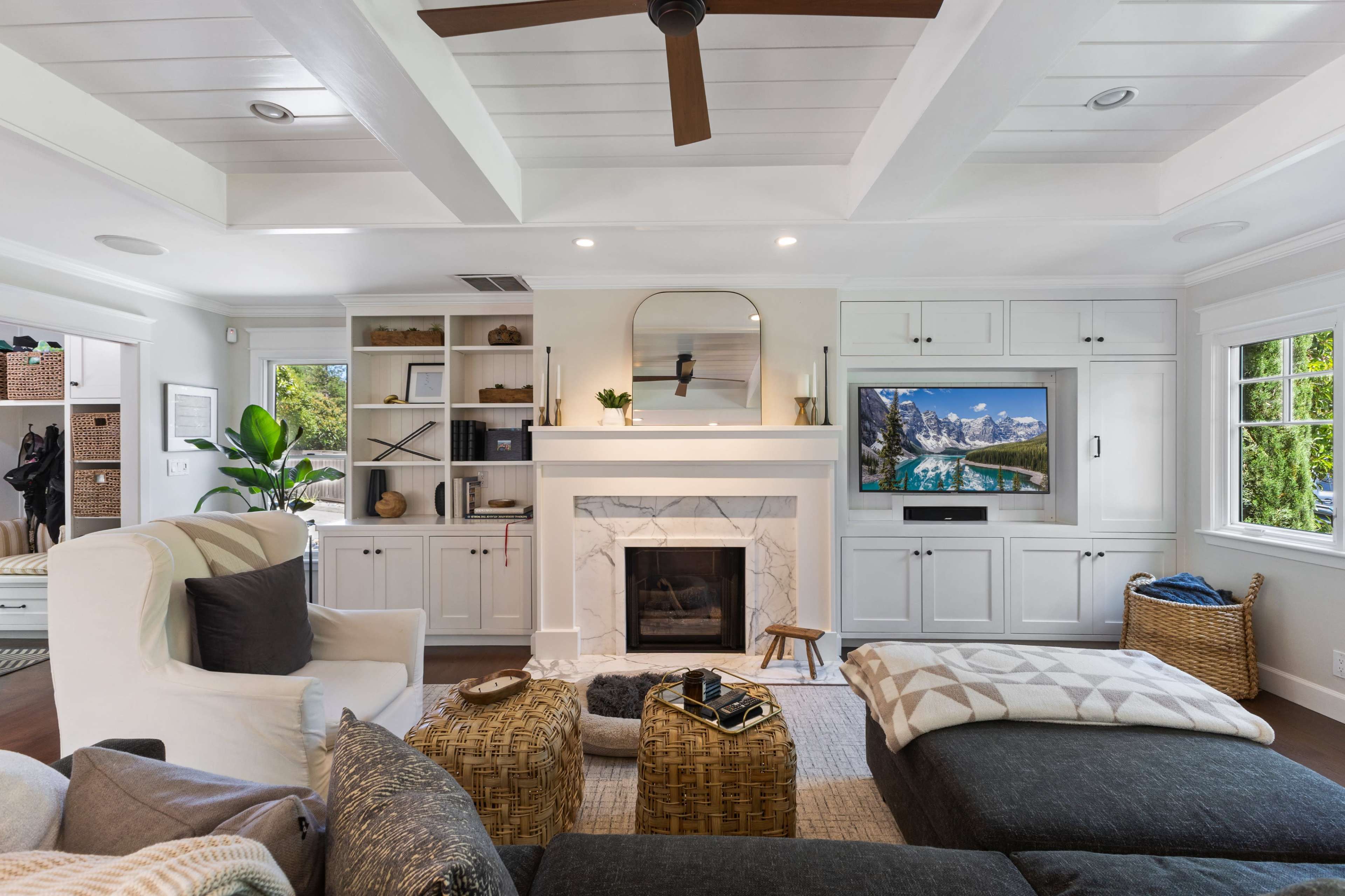 The living room features a modern design with a white fireplace, built-in shelves, a large television, and several seating options, including a sectional sofa and woven ottomans.